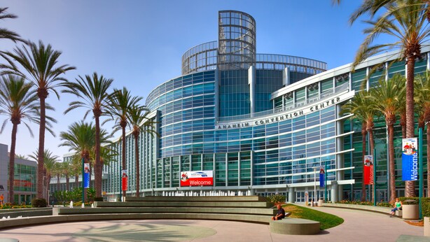 A modern building with a curved glass facade, palm trees, and a 'Welcome' sign. - Cortona Inn & Suites Anaheim Resort (Anaheim)