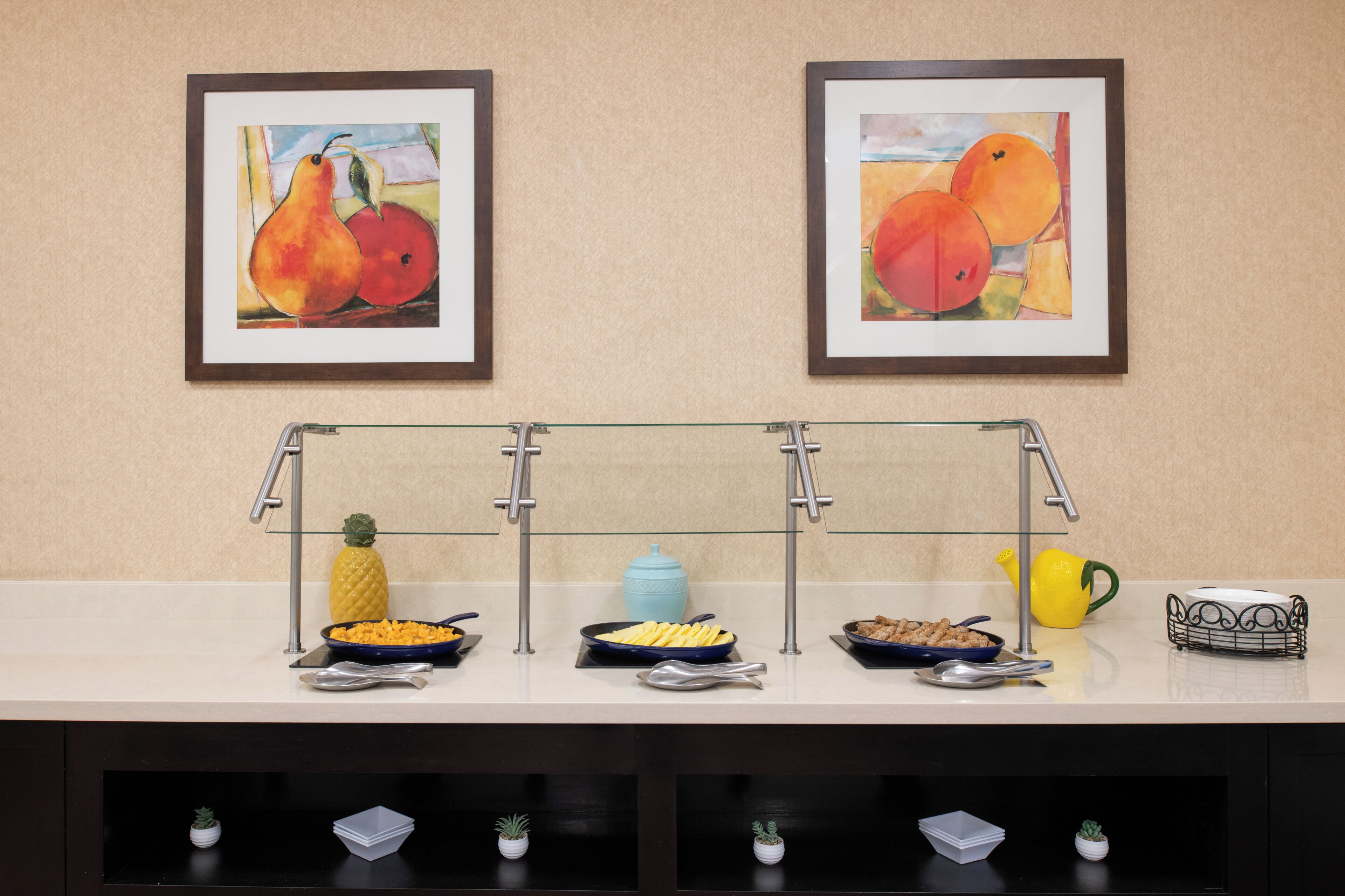 Un buffet composé de trois étagères en verre, d’un ananas, d’un pot bleu, d’un arrosoir jaune et d’un panier en fil de fer.