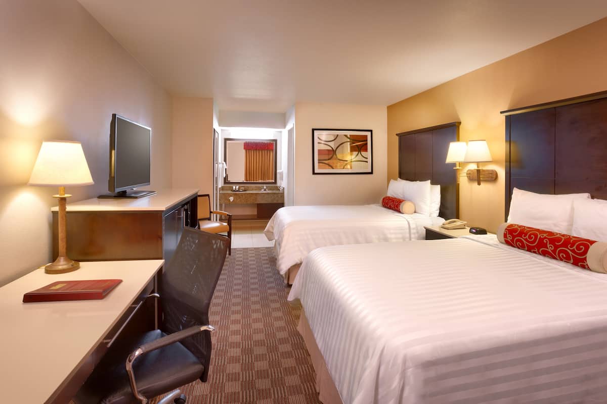 A hotel room with two beds, a desk with a TV, a chair, a desk lamp, and a bathroom visible in the background.