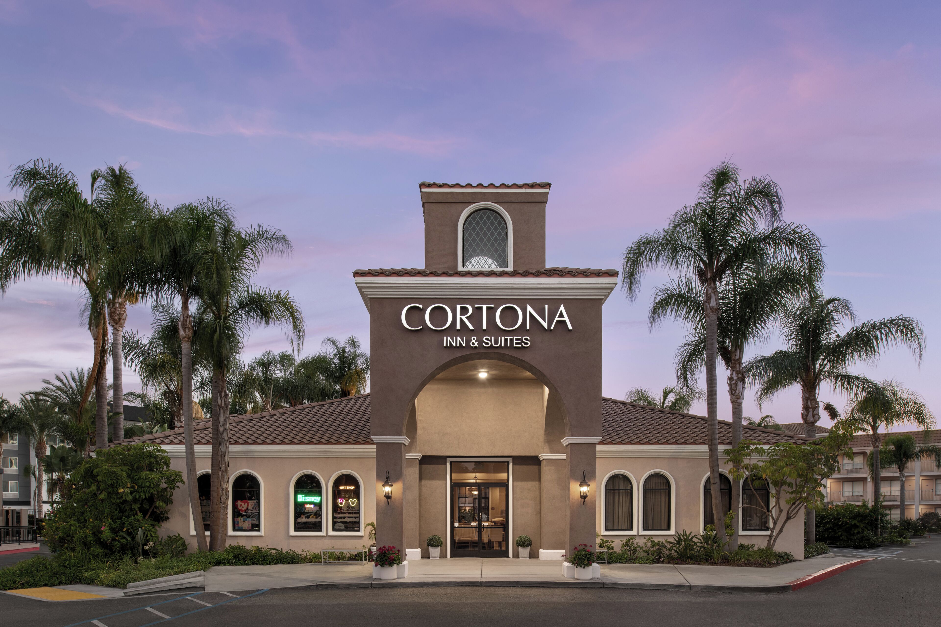A hotel named Cortona Inn & Suites with a traditional architectural style, featuring a central entrance, arched windows, and a bell tower.