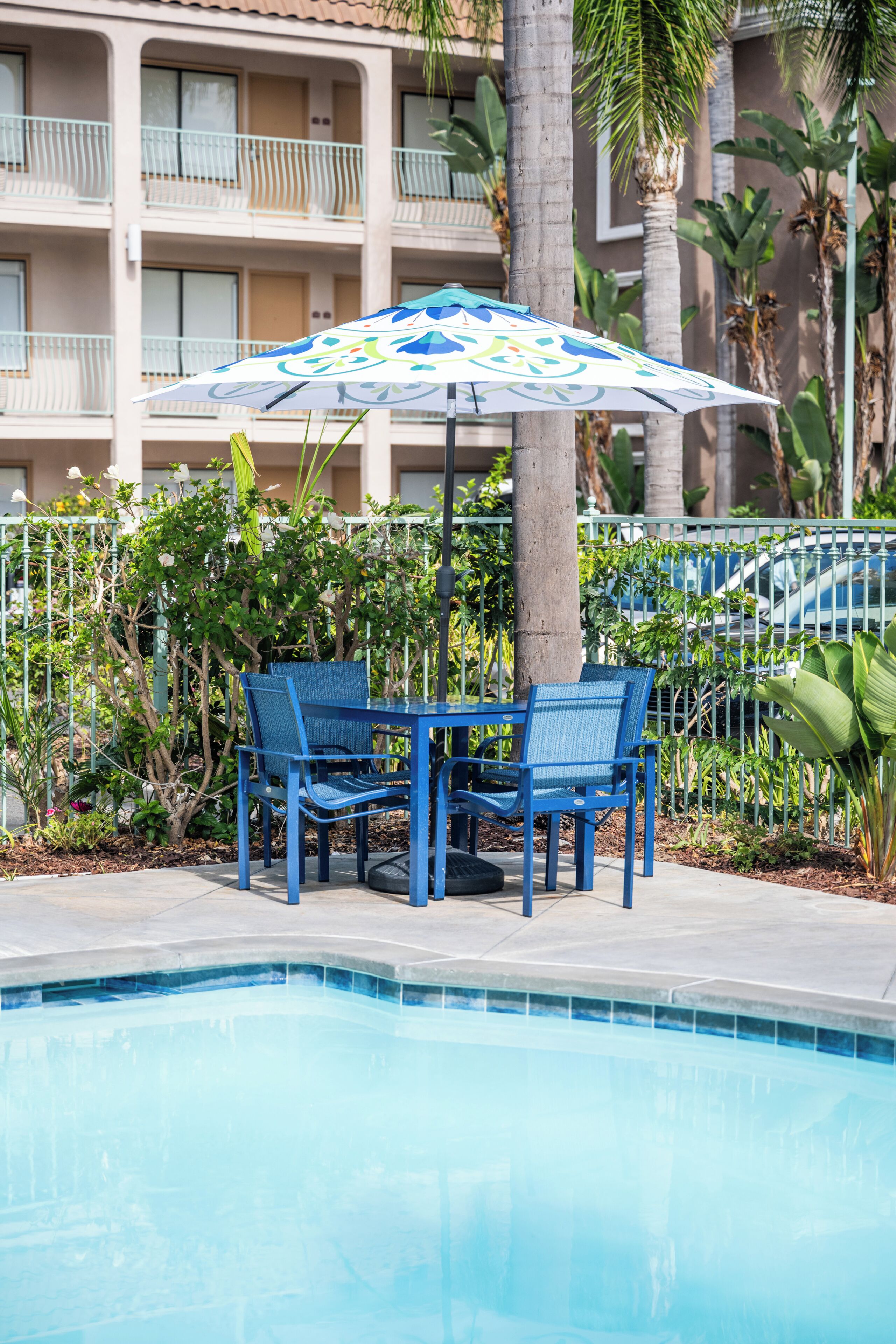 A pool area with two blue chairs and a patterned umbrella, surrounded by a fence and greenery.