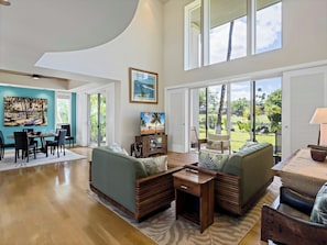 A spacious living room with a dining area, large windows, and a view of a tropical landscape.