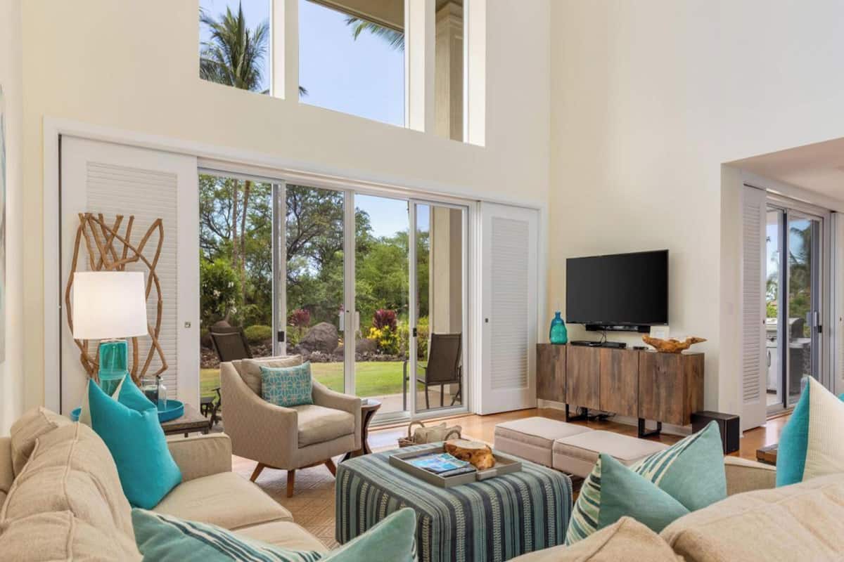A spacious living room with a large TV, comfortable seating, and a view of a lush garden through sliding glass doors.
