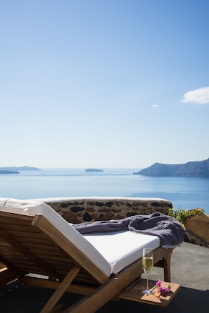 A wooden lounge chair with a white cushion and a purple towel, a glass of white wine, and a purple flower on a wooden tray. - Perivolas (Santorini)