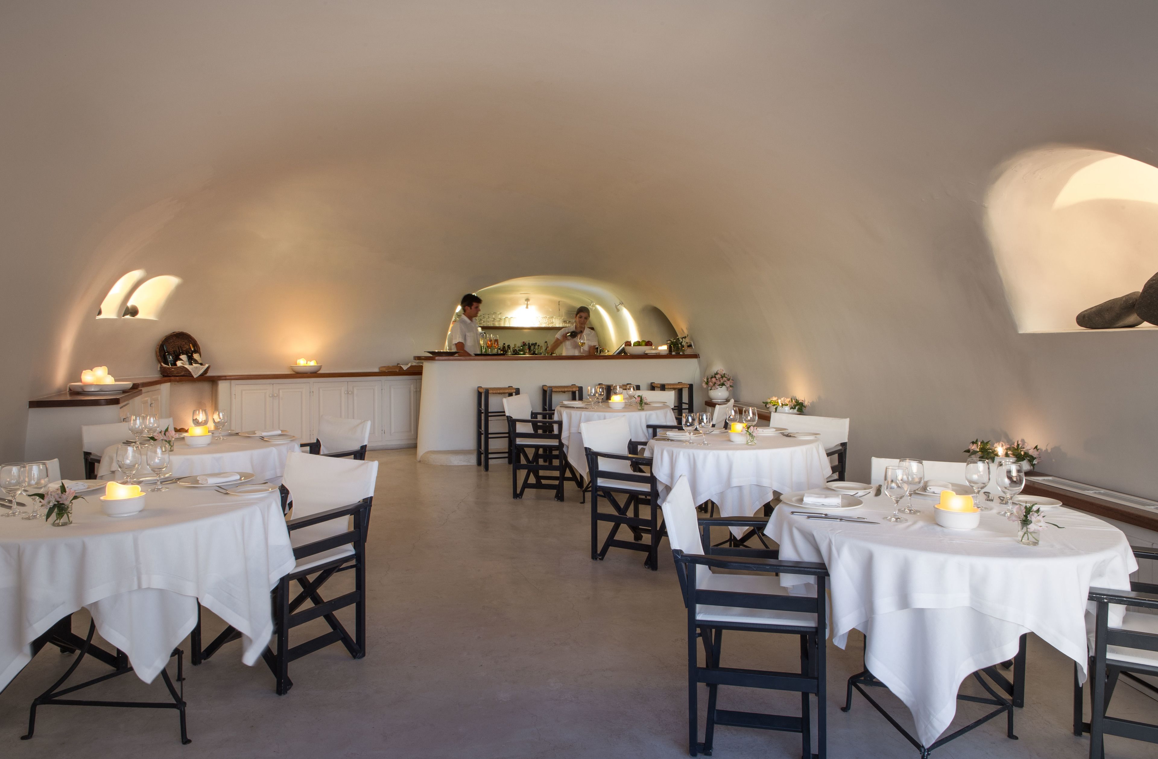 A restaurant with round tables, white tablecloths, and candles. There are chairs around the tables, and a counter with a person behind it.