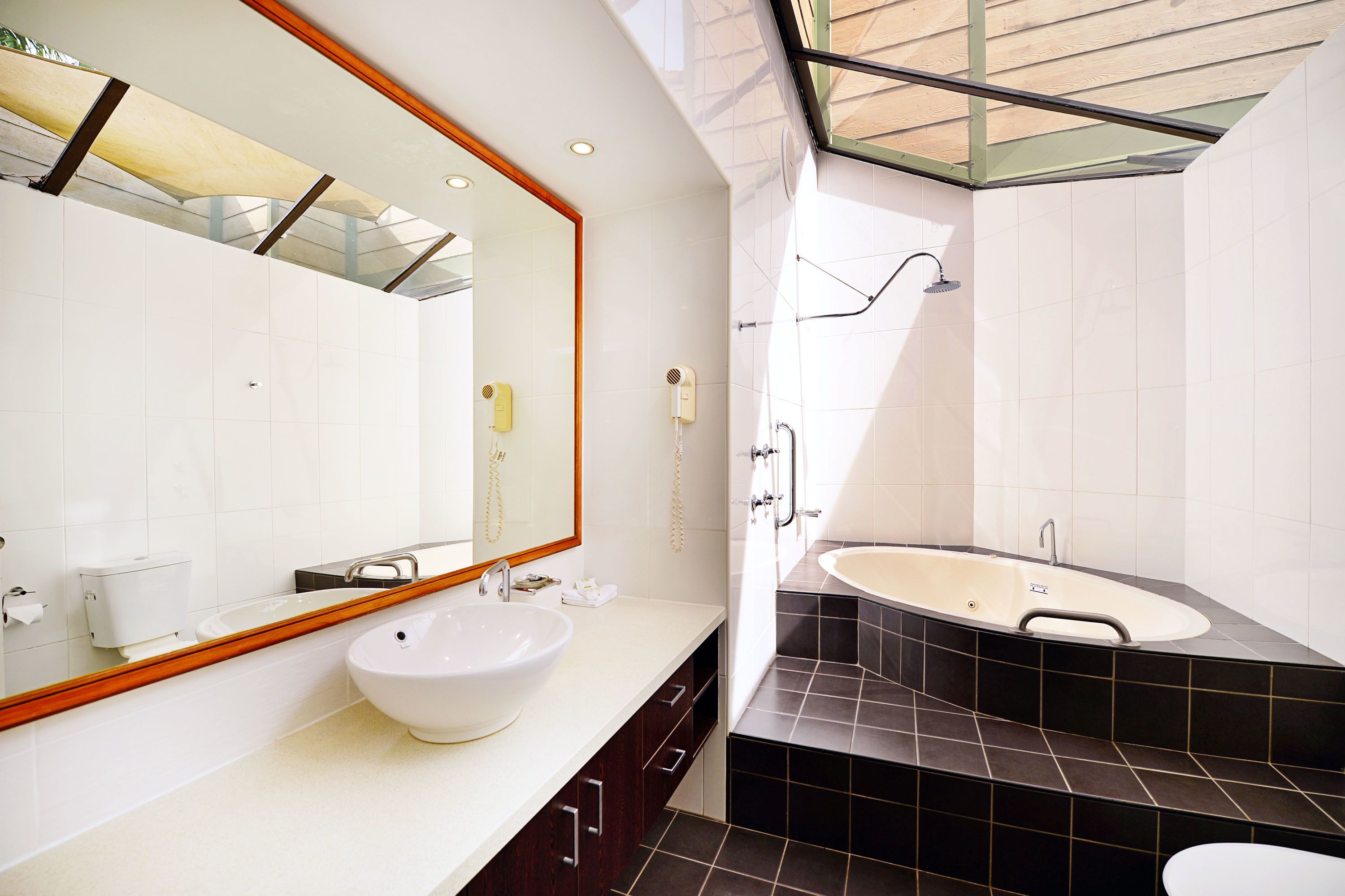 Un baño moderno con un espejo grande, un lavabo blanco, una bañera y suelo de baldosas oscuras.