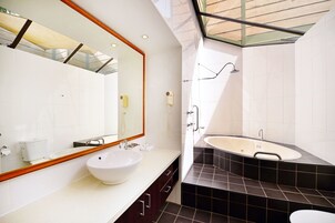 Un baño moderno con un espejo grande, un lavabo blanco, una bañera y suelo de baldosas oscuras.