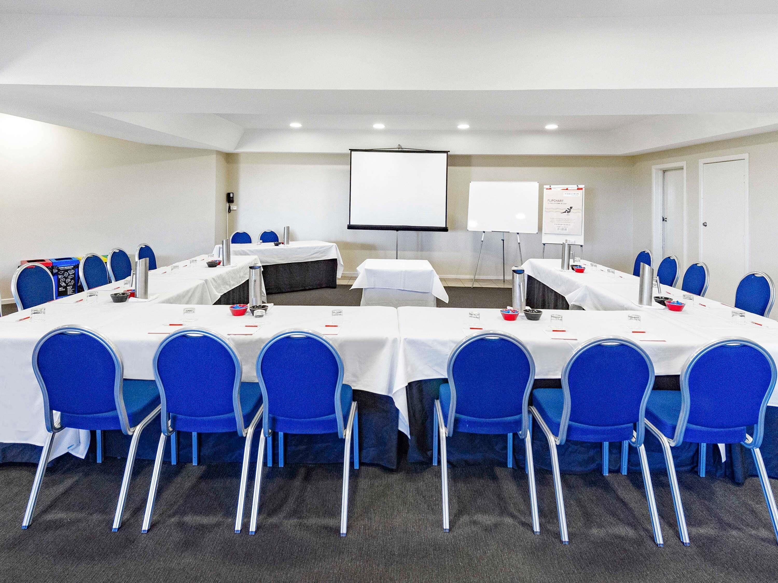 Une salle de conférence avec une longue table, des chaises bleues, des plaques nominatives et un écran de projection.