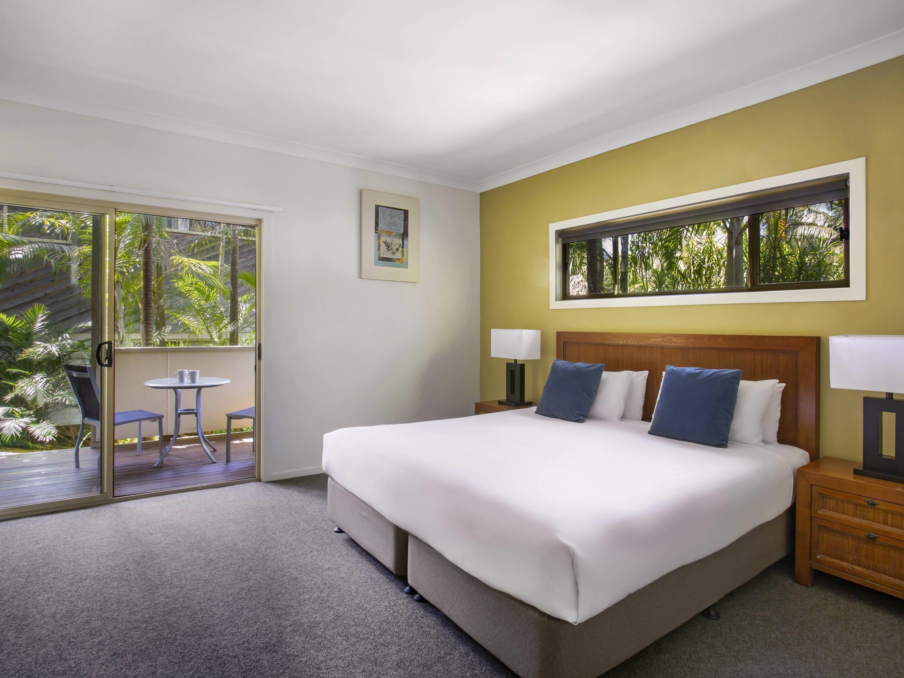 Une chambre d’hôtel comprenant un lit, deux tables de chevet avec des lampes, une fenêtre donnant sur un espace verdoyant, et une porte-fenêtre coulissante qui mène à un espace extérieur avec une table et des chaises.