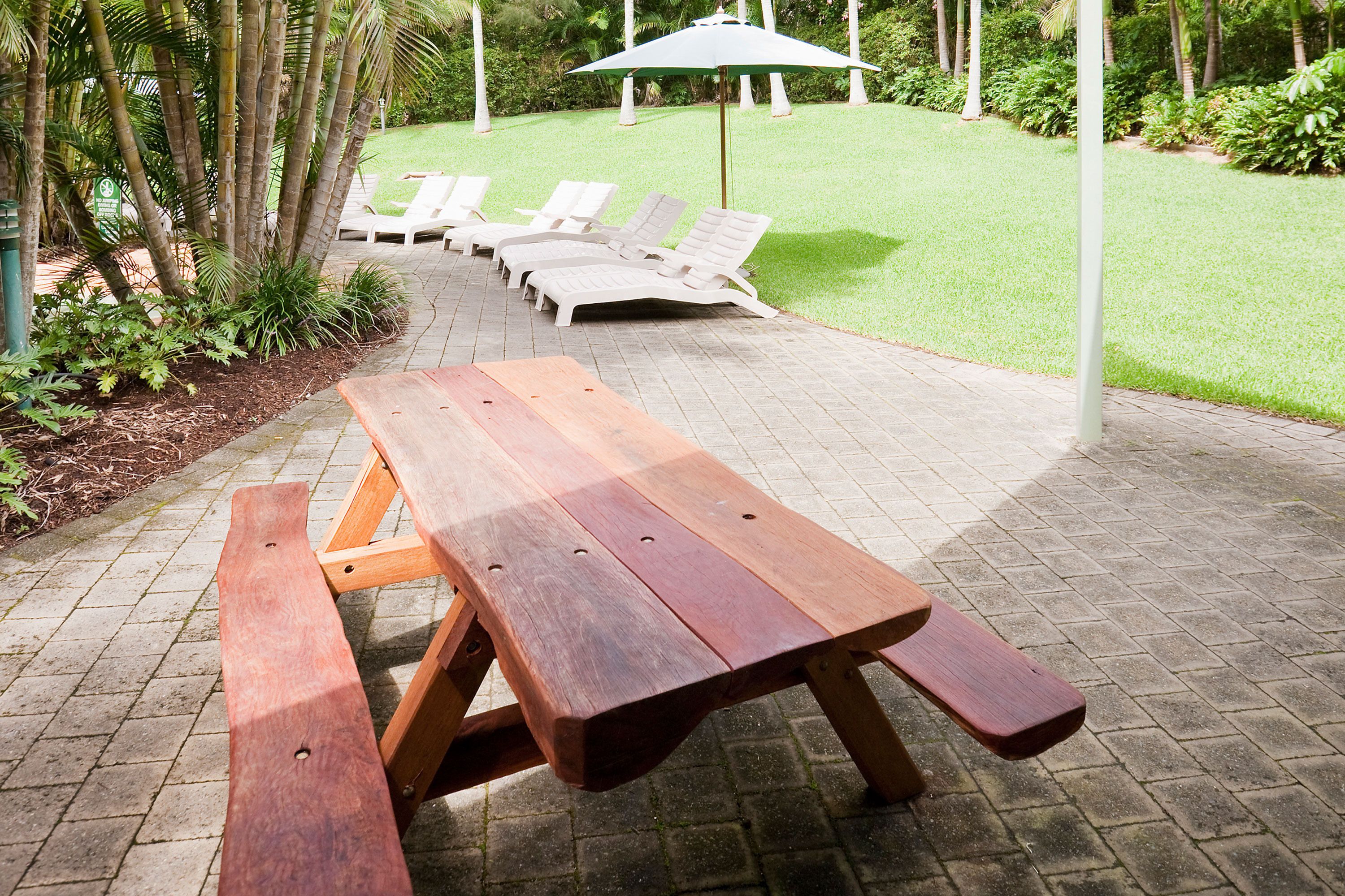 A wooden picnic table with benches, surrounded by a grassy area with white lounge chairs and a green umbrella.