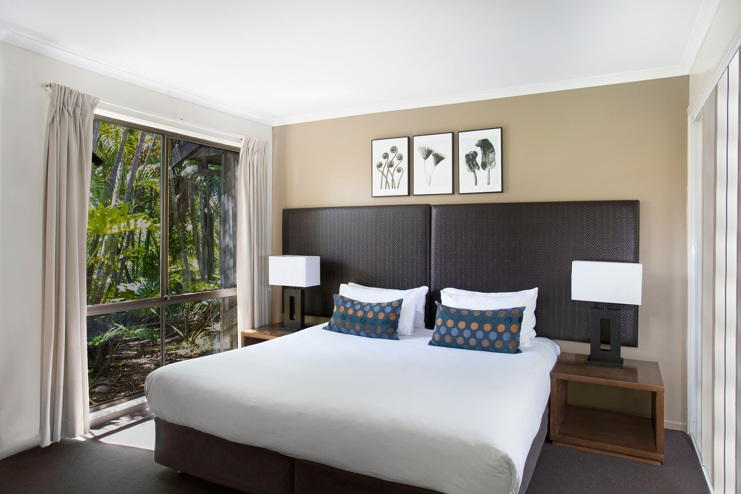 Una habitación de hotel con una cama grande, dos mesitas de noche con lámparas, una ventana con cortinas y tres cuadros en la pared.