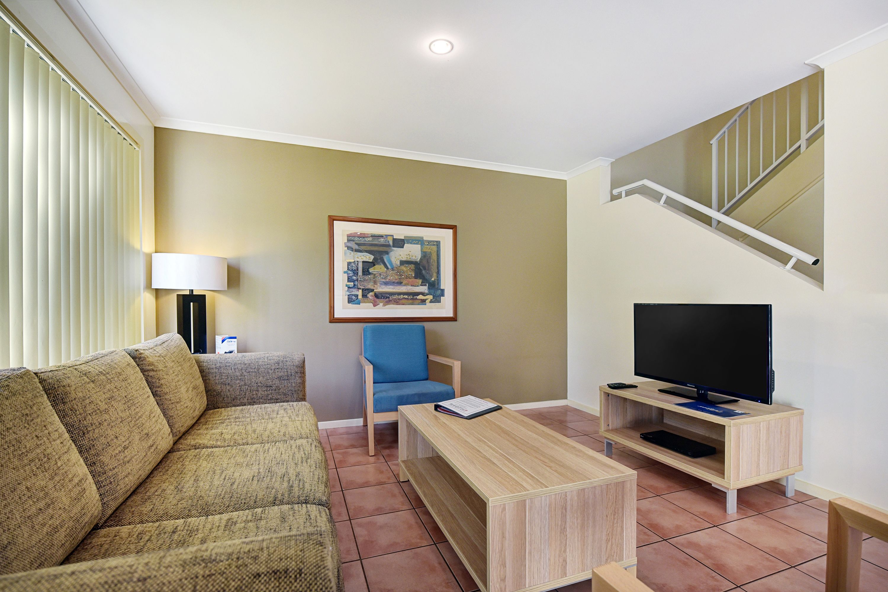 Una sala de estar con un sofá, una silla, una mesita de centro, un televisor sobre un mueble bajo, un cuadro en la pared y una escalera.