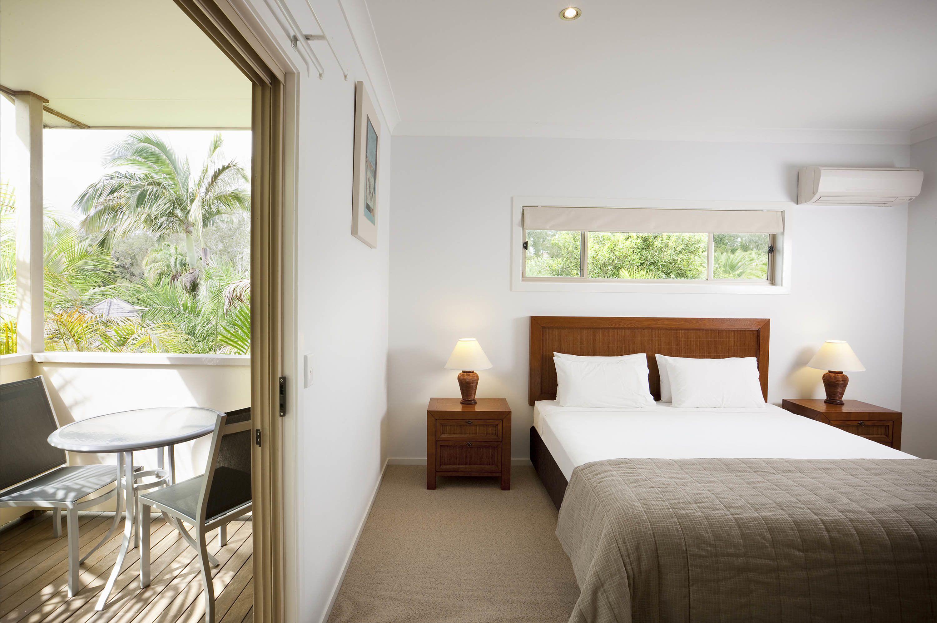 A hotel room with a bed, two bedside tables with lamps, a small table with chairs, and a window with a view of greenery.