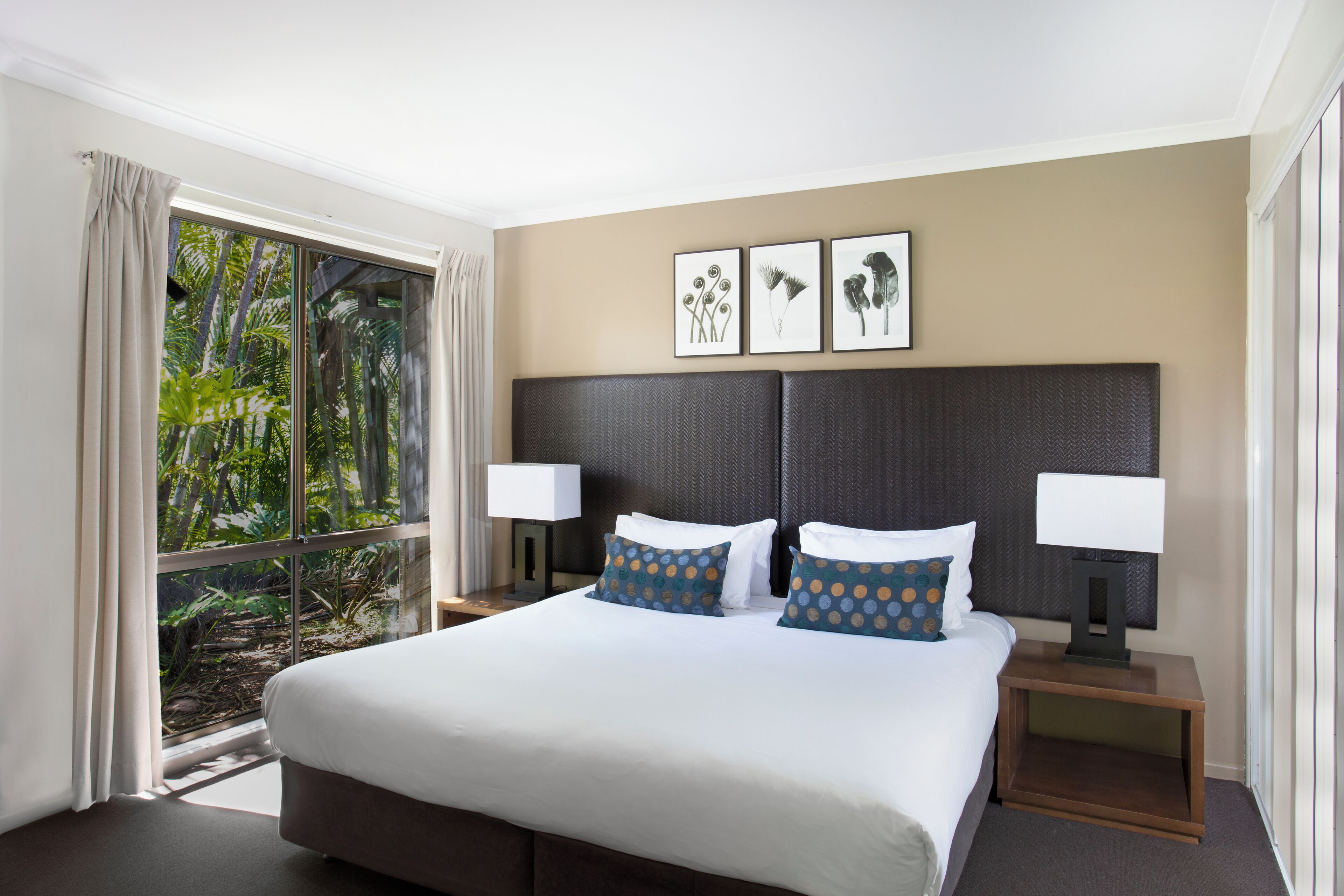 Une chambre d’hôtel avec un grand lit, deux tables de chevet avec des lampes, une fenêtre avec des rideaux et trois tableaux encadrés accrochés au mur.