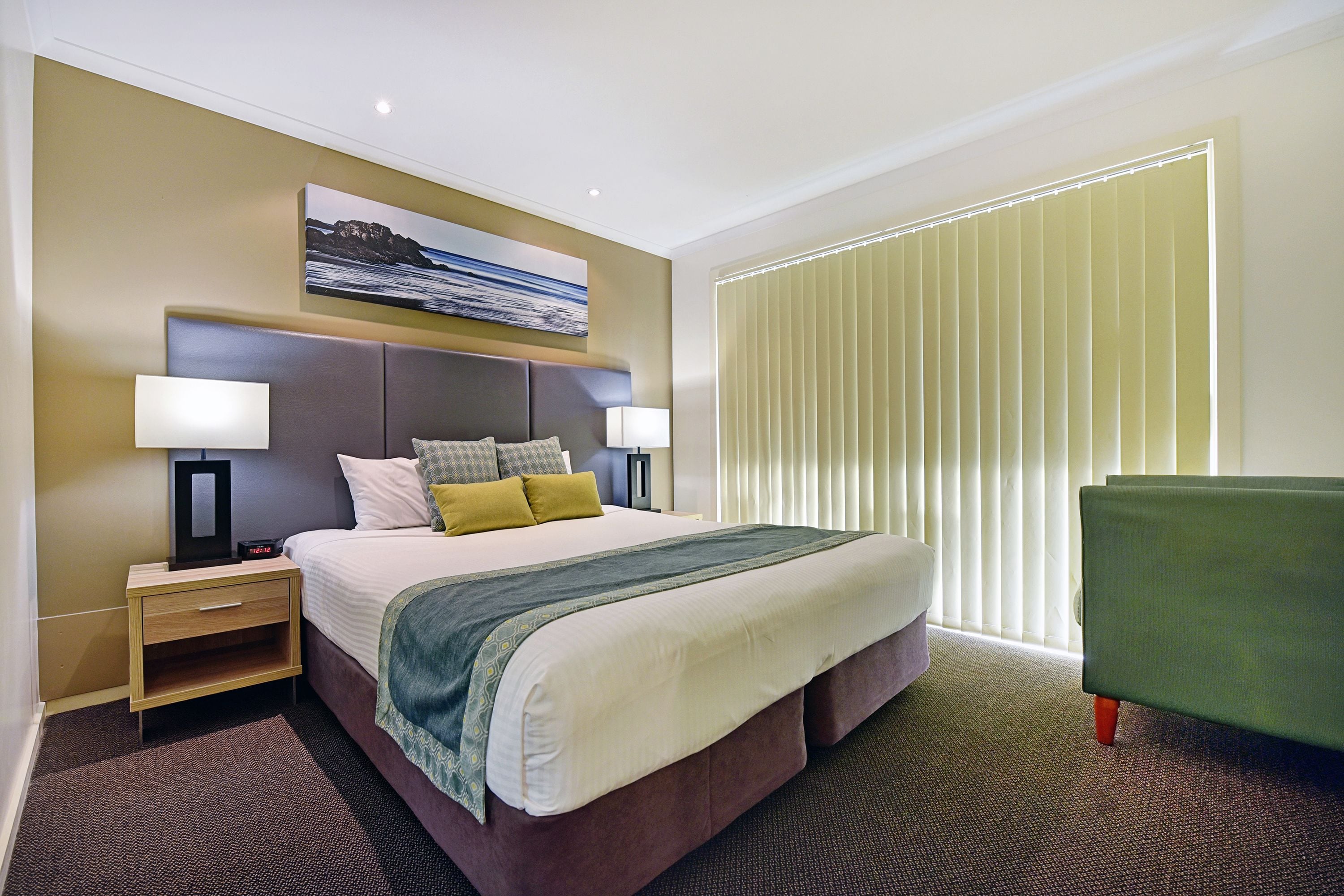 A hotel room with a large bed, two bedside tables, a nightstand, a green armchair, and a window with blinds.