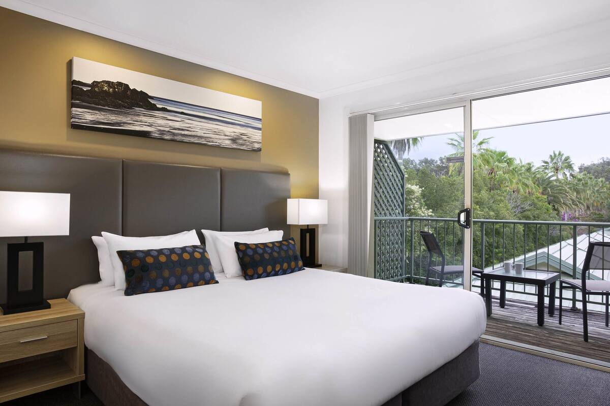 Ein Hotelzimmer mit einem großen Bett, zwei Nachttischlampen, einem Gemälde über dem Bett und einem Balkon mit Blick auf Bäume und ein Gebäude.