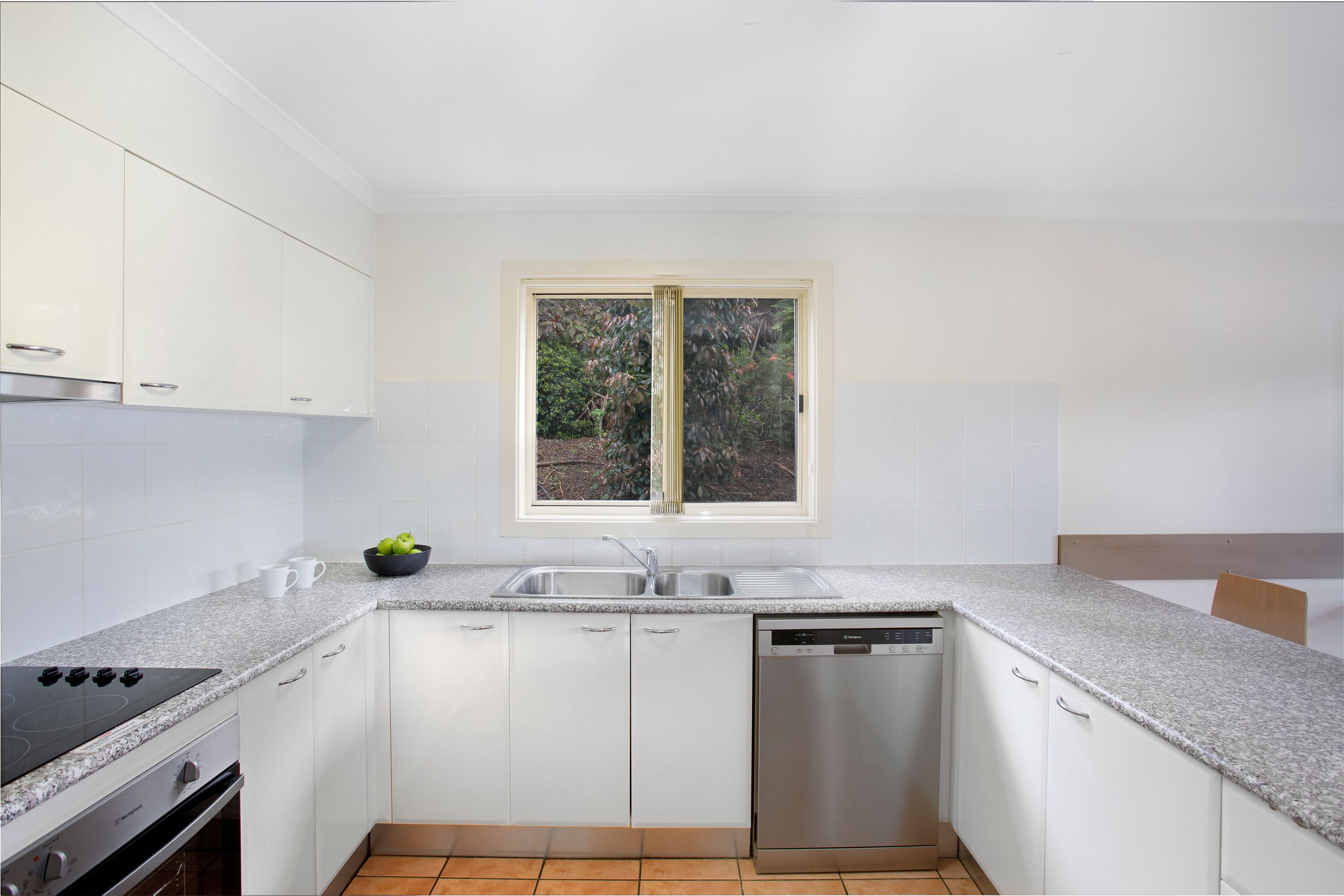 A modern kitchen with white cabinets, a built-in stove, a dishwasher, and a window with a view of greenery.
