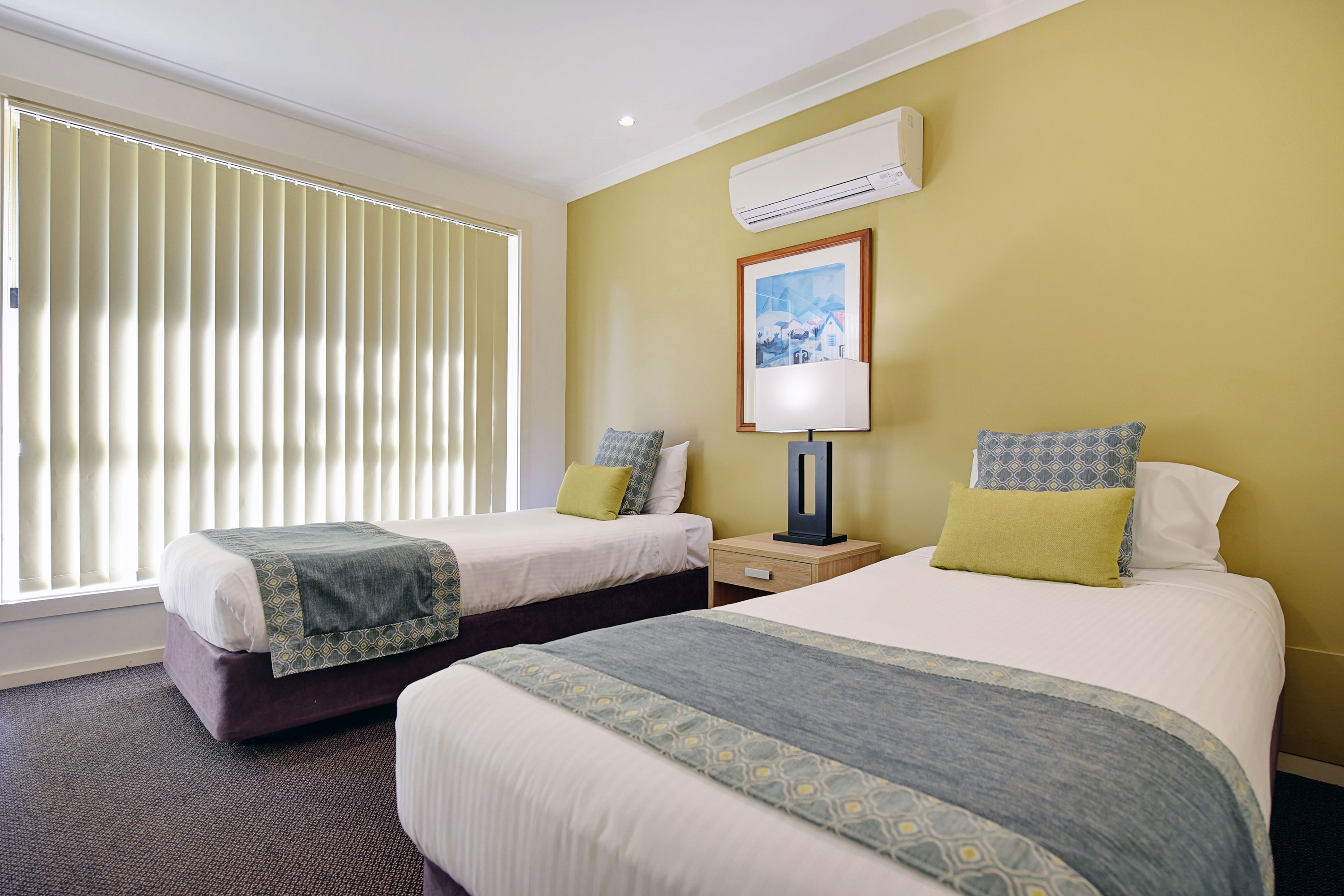 Une chambre d’hôtel avec deux lits, une table de chevet, une lampe, un tableau encadré au mur et un appareil de climatisation.