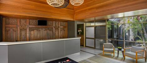 A reception area with a wooden ceiling, a reception desk, two chairs, a small table with a bowl, and a large window with a view of greenery.