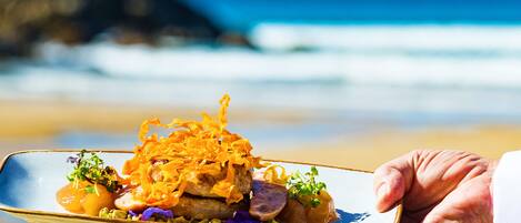 A hand holding a plate with food, set against a beach and ocean backdrop.