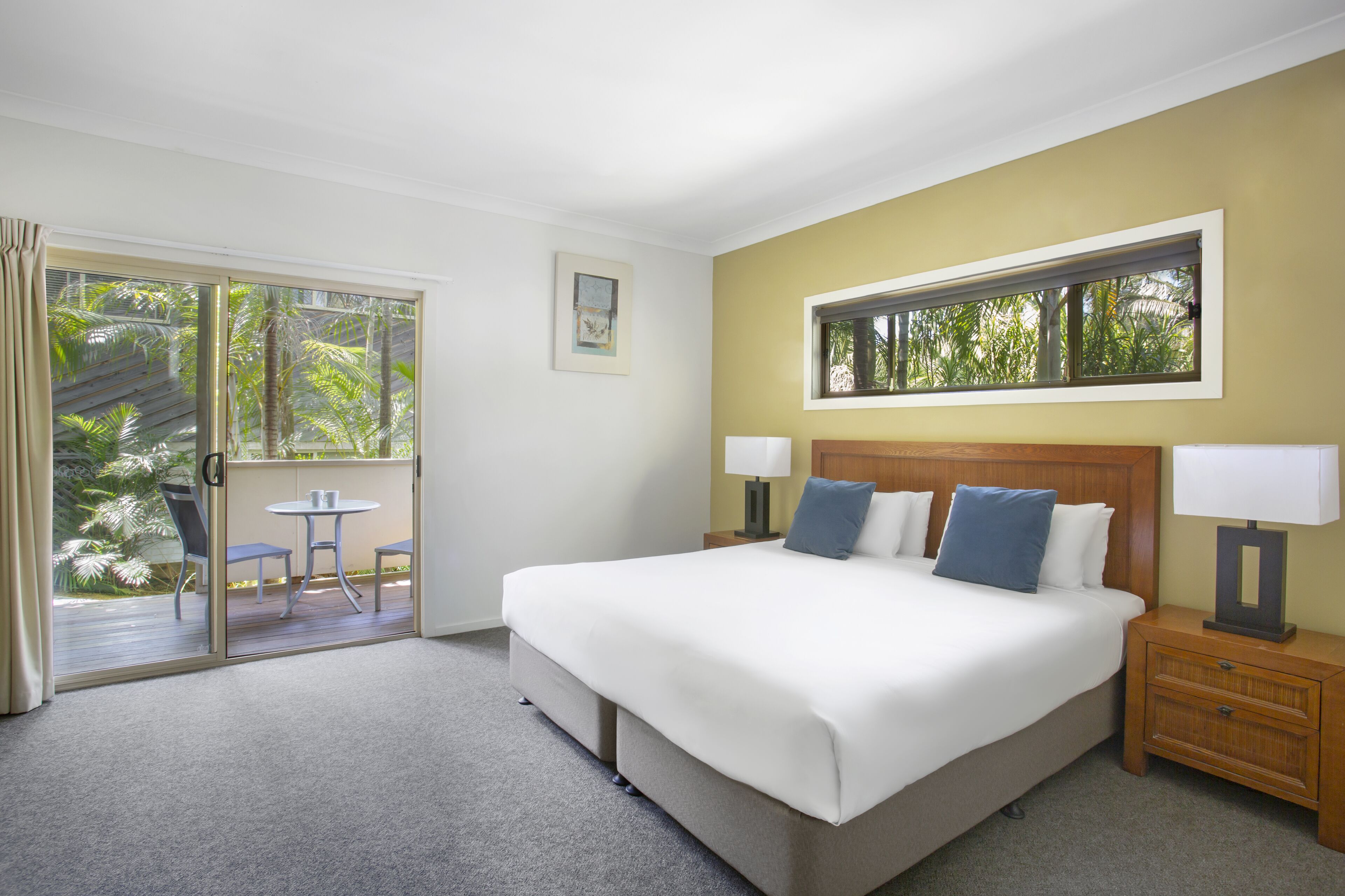 Une chambre d’hôtel avec un grand lit, une petite table, deux chaises, une fenêtre donnant sur des palmiers et une table de chevet en bois avec une lampe.