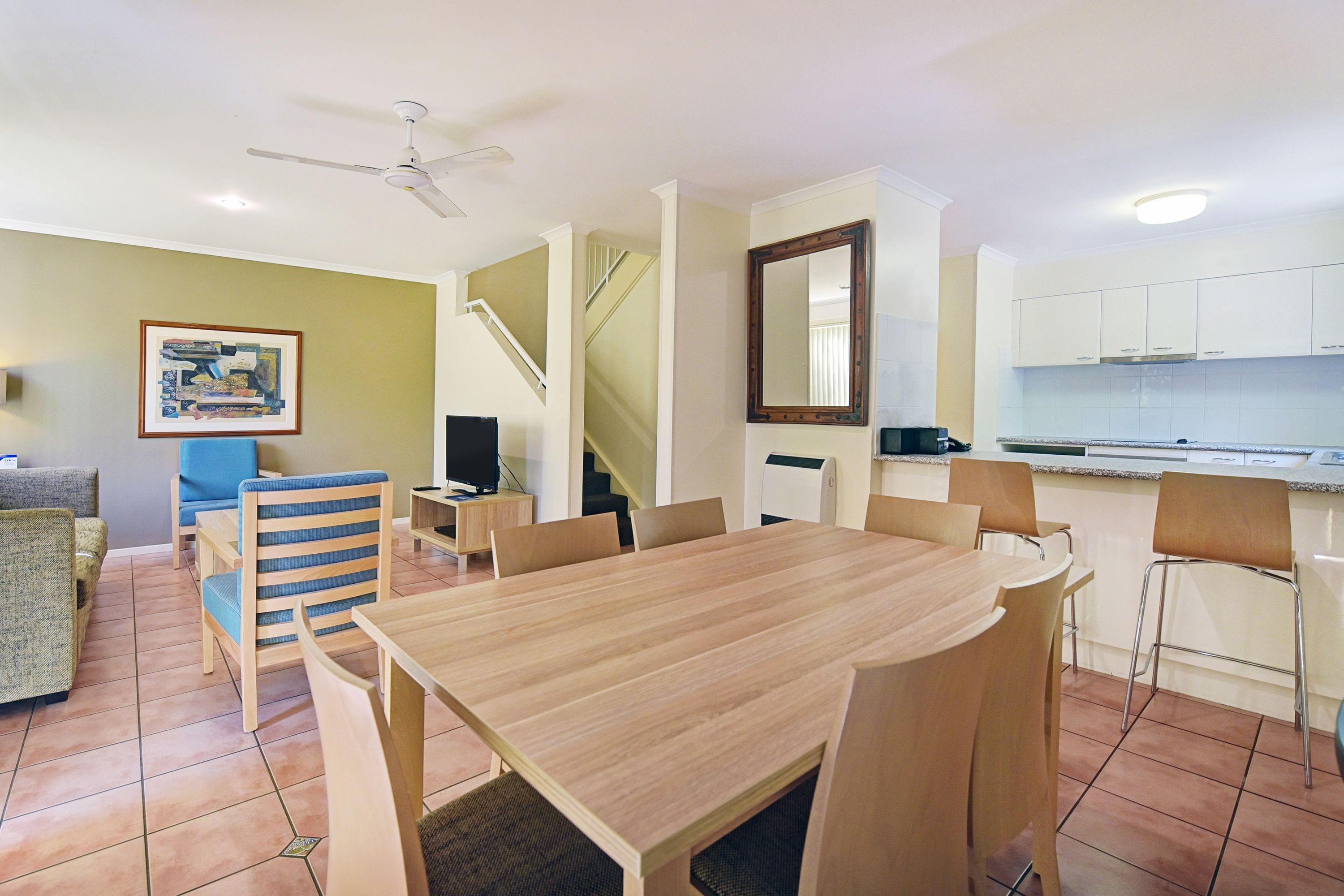 A dining area with a wooden table and chairs, a staircase, a kitchen area, and a framed picture on the wall.