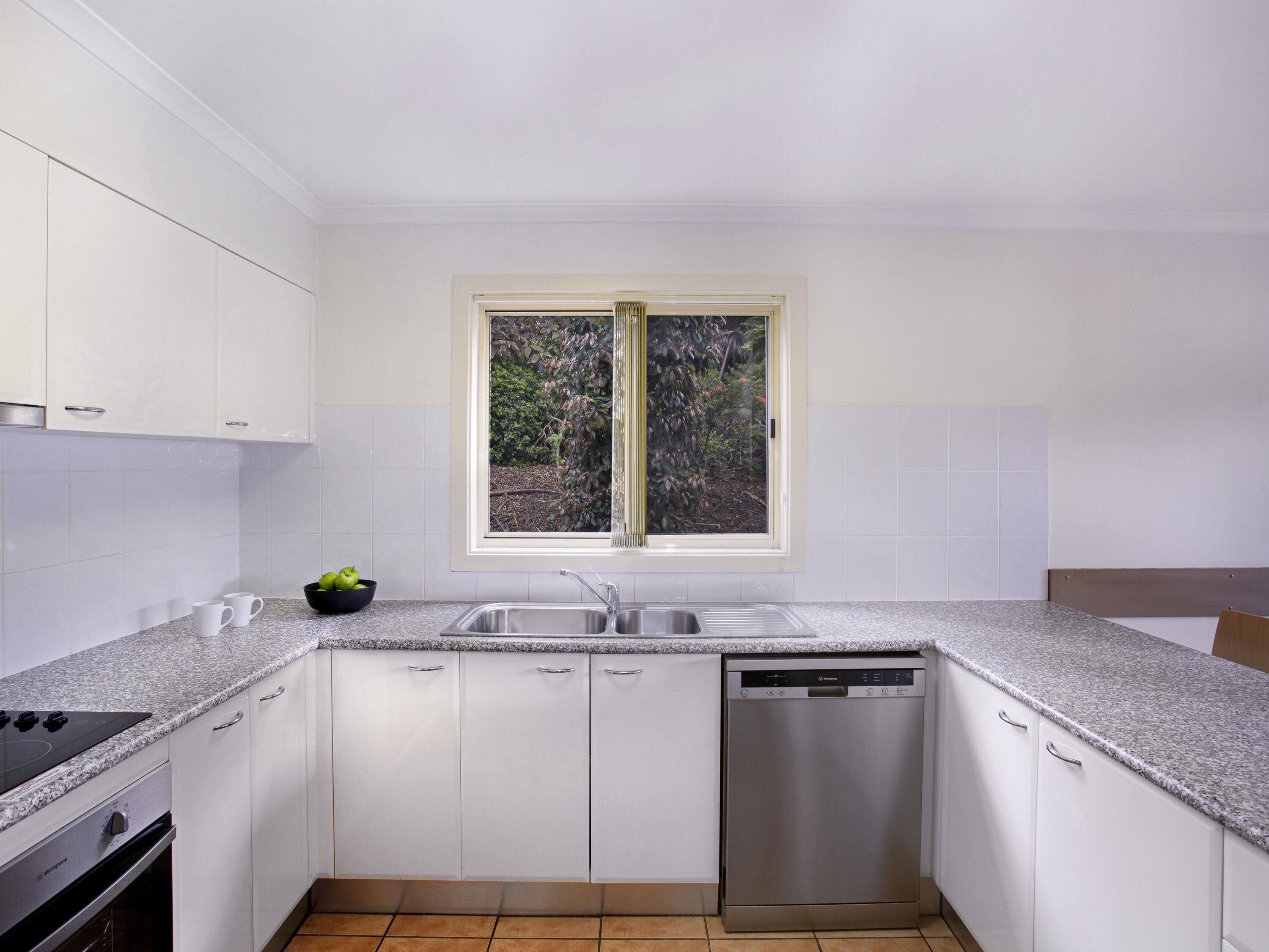 A modern kitchen with white cabinets, a stainless steel dishwasher, and a window with a view of greenery.