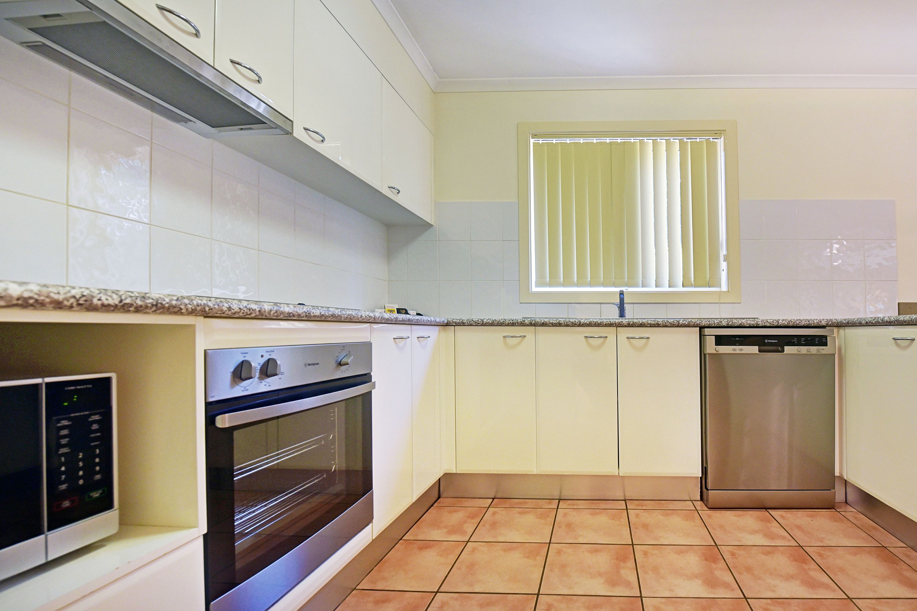 Una cocina con gabinetes blancos, horno empotrado, lavavajillas y una ventana con persianas.