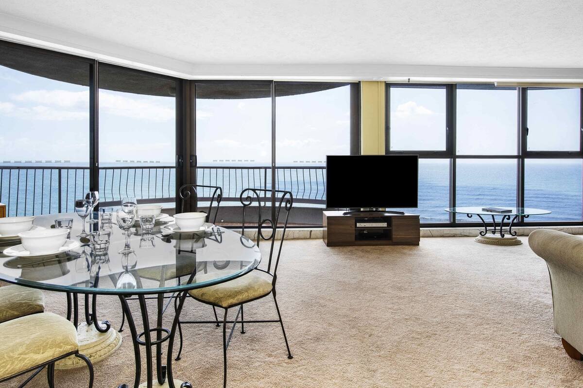 A modern living room with a glass table, chairs, a flat-screen TV, and a balcony with a view of the sea.