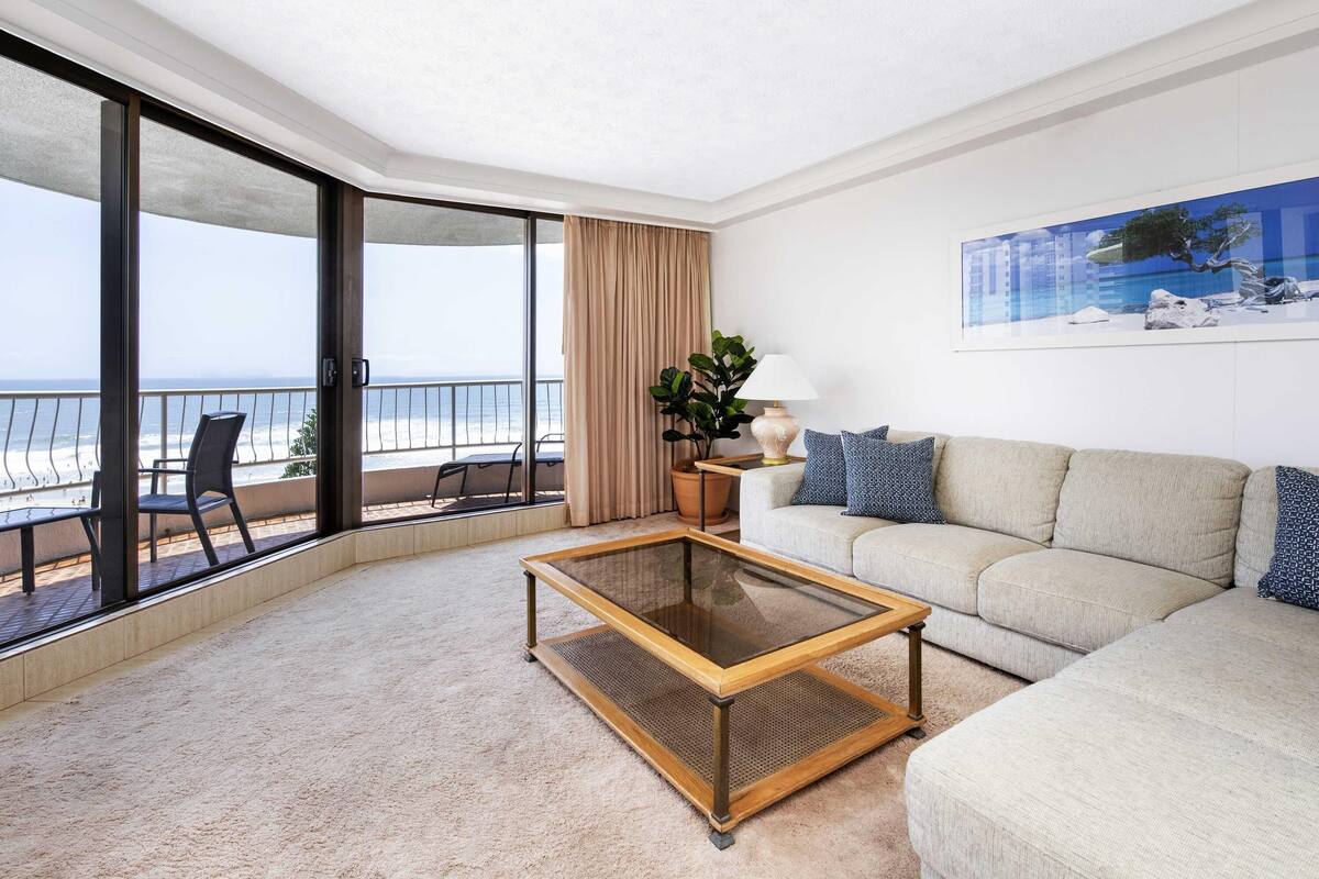 A modern living room with a glass coffee table, a beige sofa, a potted plant, and a view of the ocean.