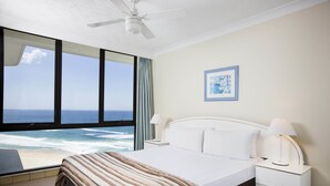 Una habitación de hotel con un ventanal grande que ofrece vistas al océano, una cama con manta a rayas, un cabecero blanco y una mesita de noche con una lámpara.