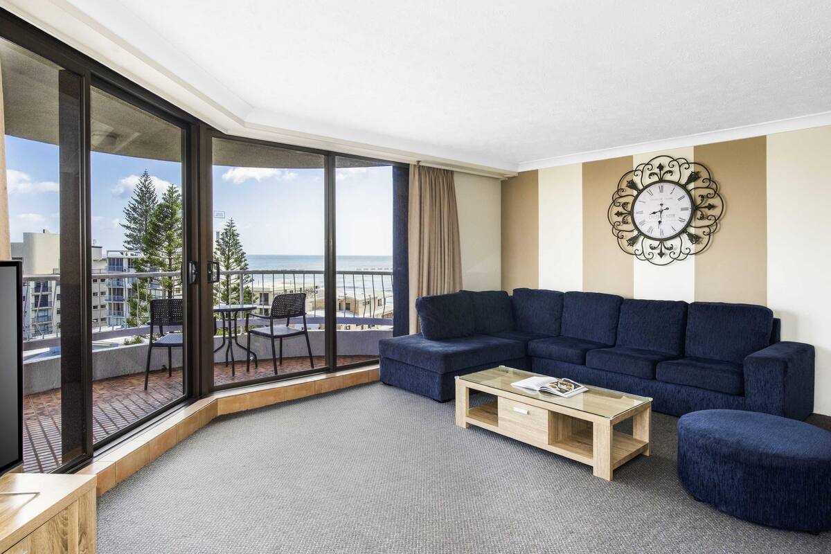 A modern living room with a large window, a blue sofa, a wooden coffee table, and a decorative wall clock.