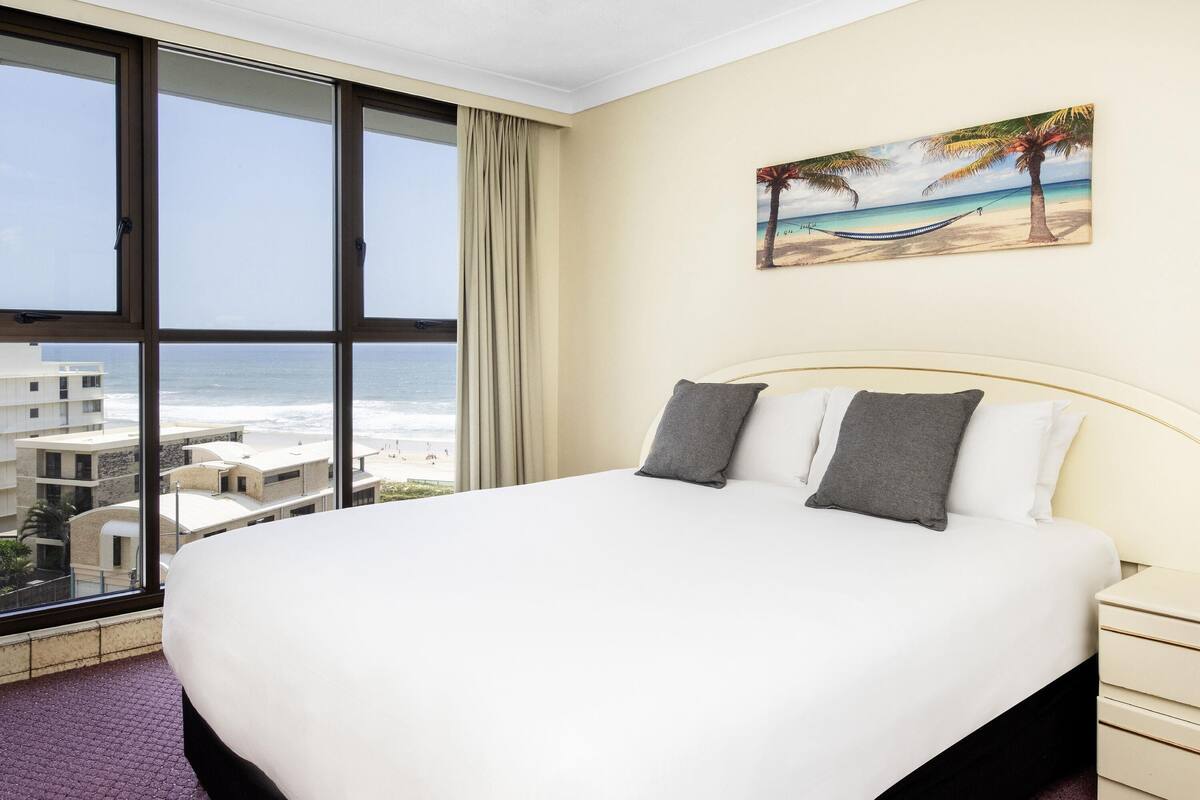 A hotel room with a large bed, a painting of a beach scene, and a view of the ocean through a large window.