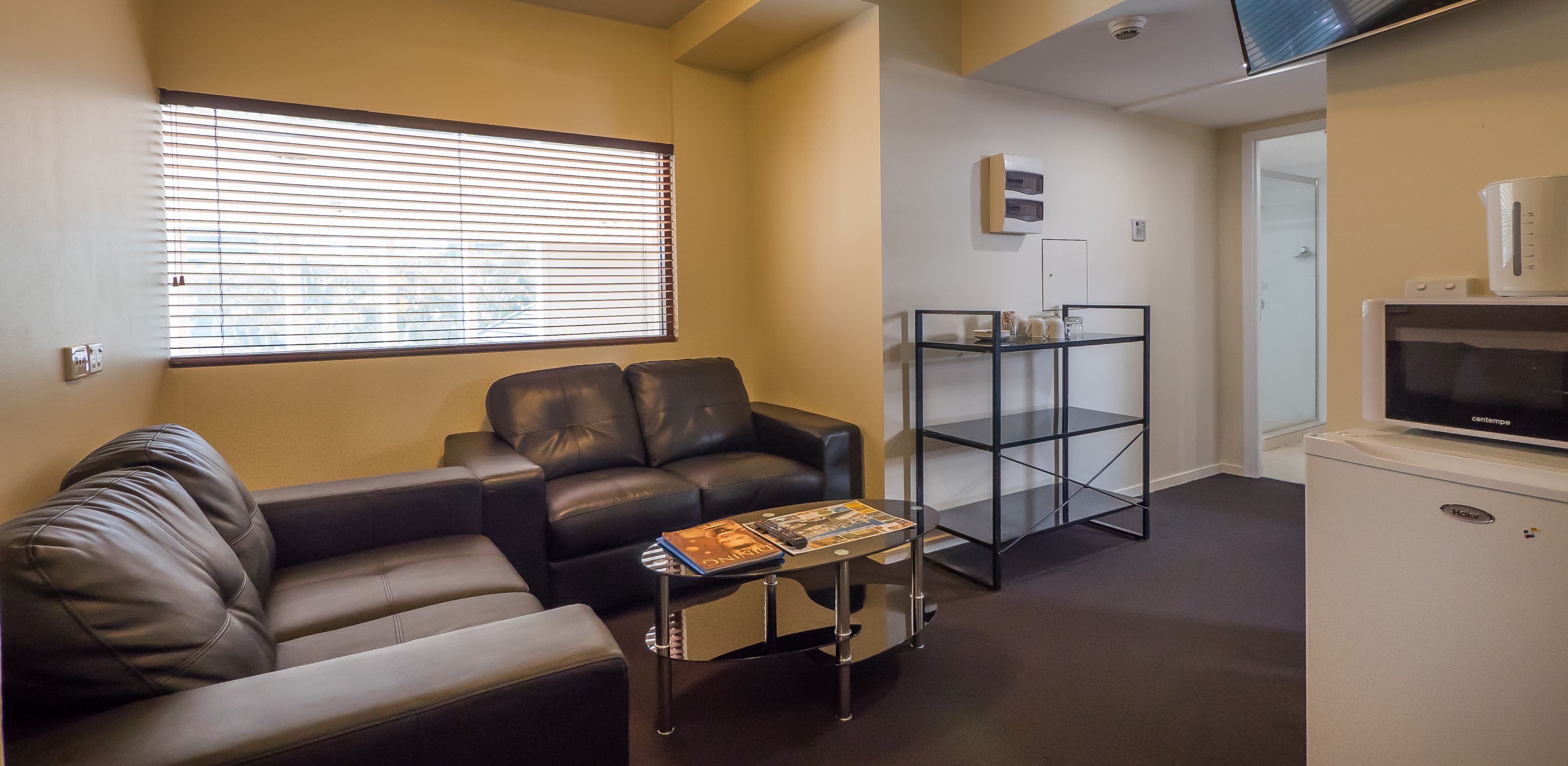 Un salon avec un canapé en cuir, une table basse en verre sur laquelle sont posés des magazines, un téléviseur sur un meuble et une petite armoire blanche.