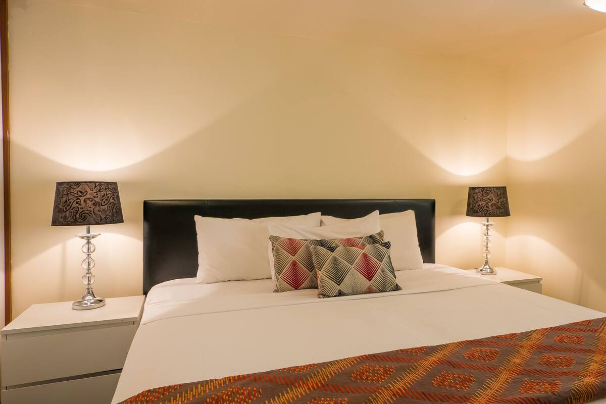 Een net opgemaakt bed met witte lakens en een decoratieve plaid, met aan beide zijden nachtkastjes en lampen, in een kamer met een effen muur en een klein nachtkastje.