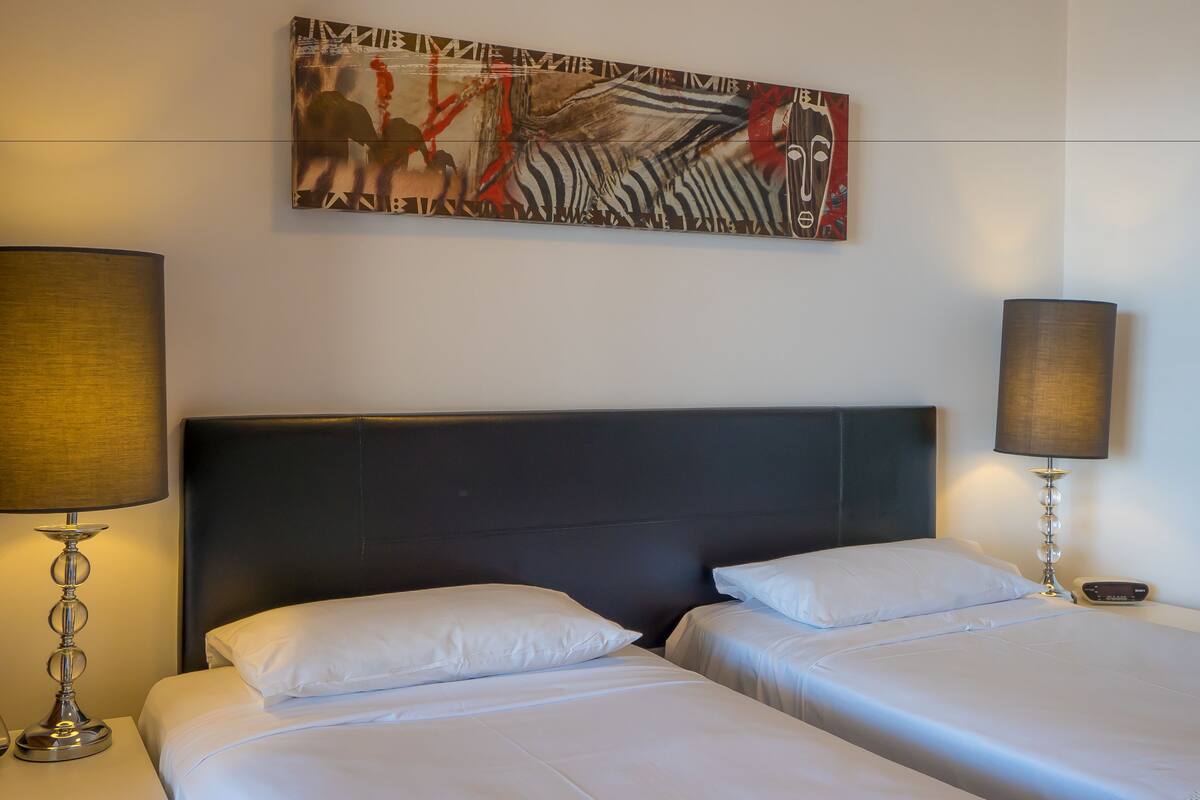 Een hotelkamer met twee bedden, een groot abstract schilderij aan de muur en lampen aan weerszijden.