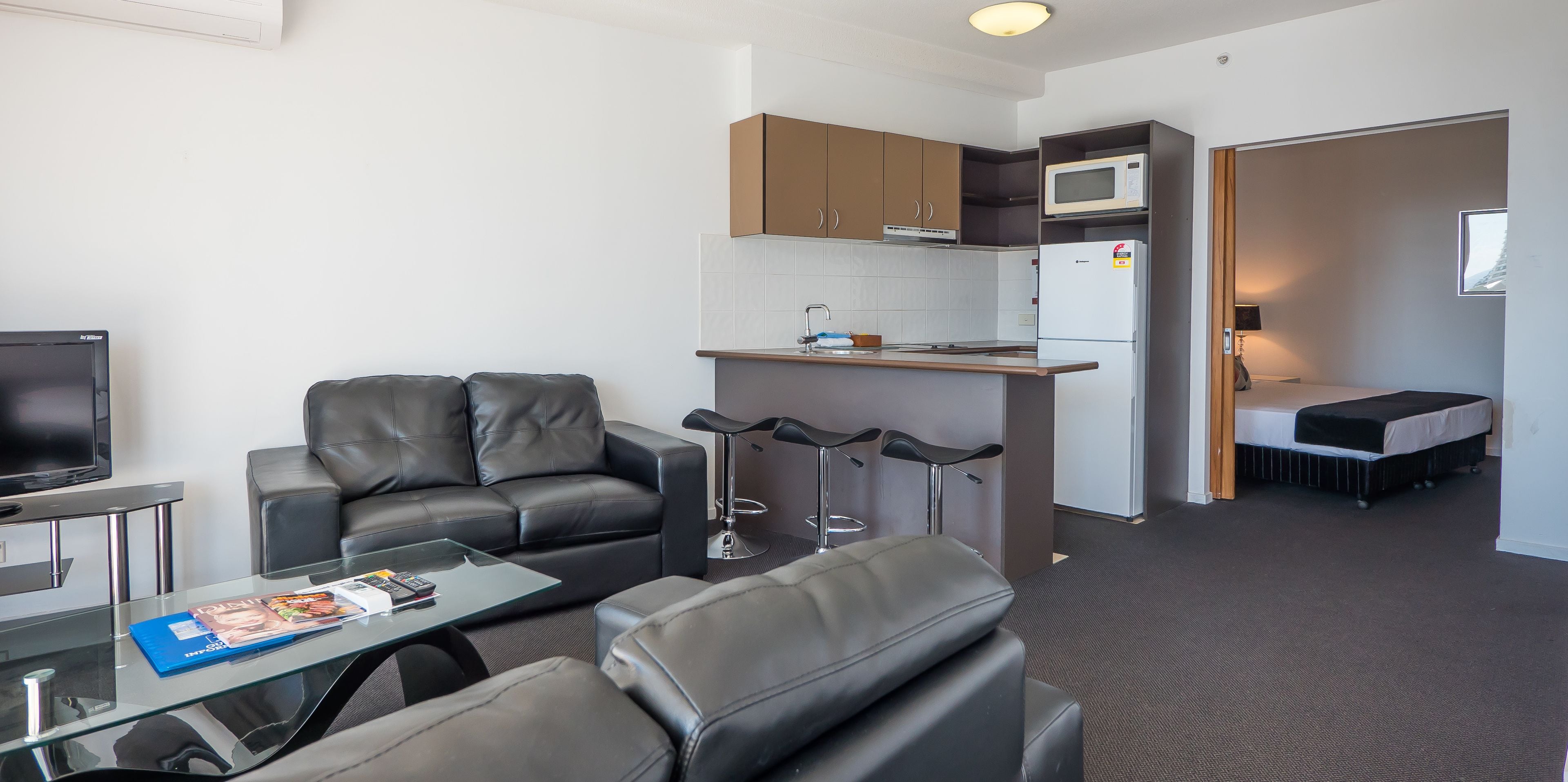 A compact living space with a sofa, a glass coffee table, a kitchenette, a small bar area, and a separate sleeping area.