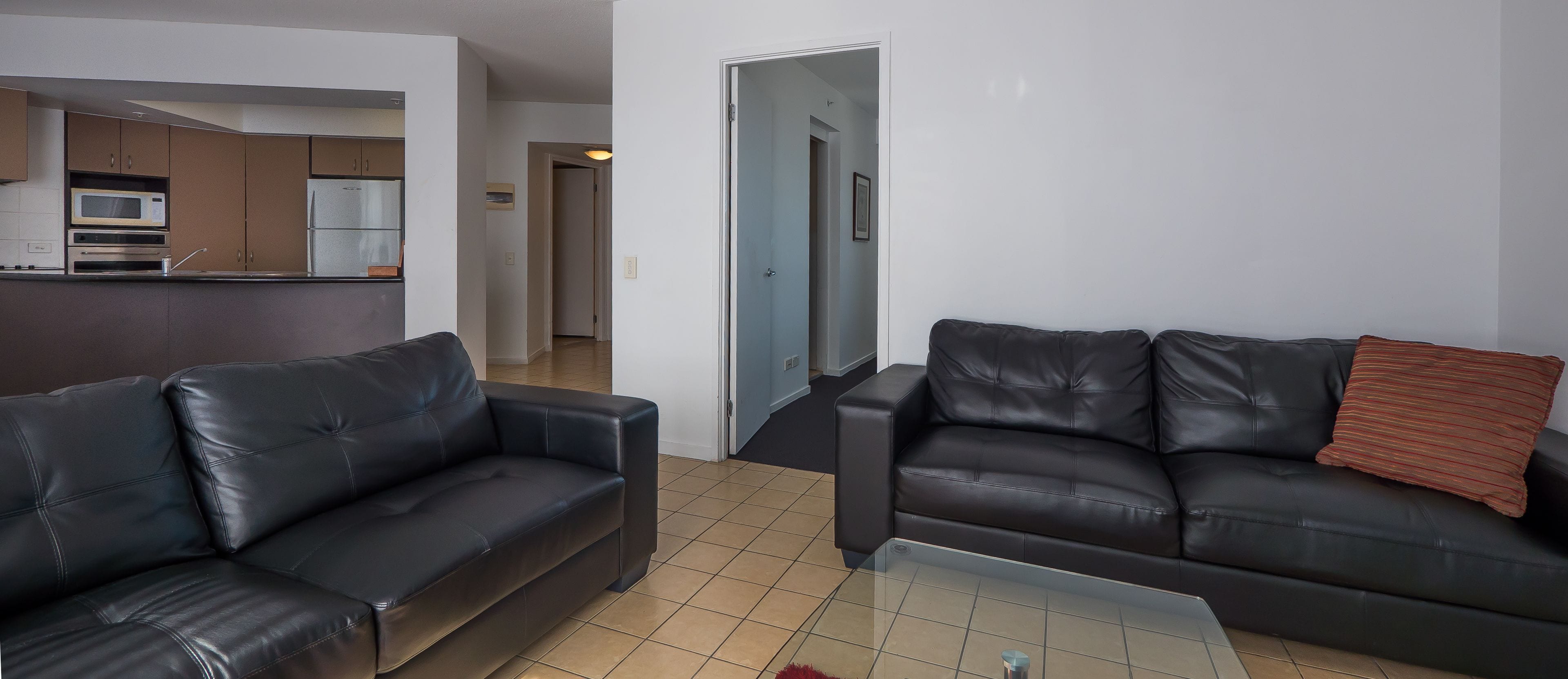 A living room with two black leather sofas, a glass coffee table, and a kitchen area with a microwave and refrigerator.