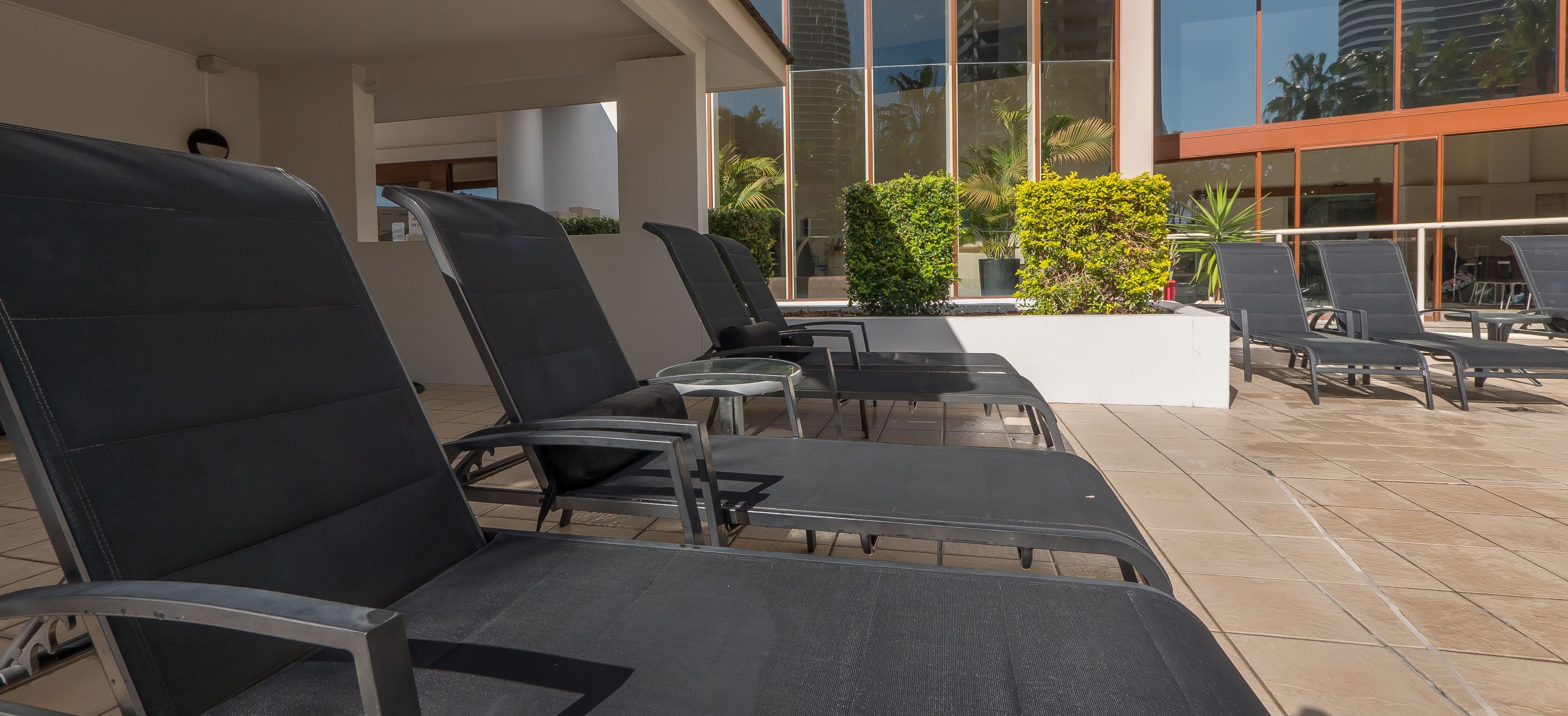 Une rangée de fauteuils noirs et une petite table sont disposées sur une terrasse carrelée, avec un bâtiment en arrière-plan.
