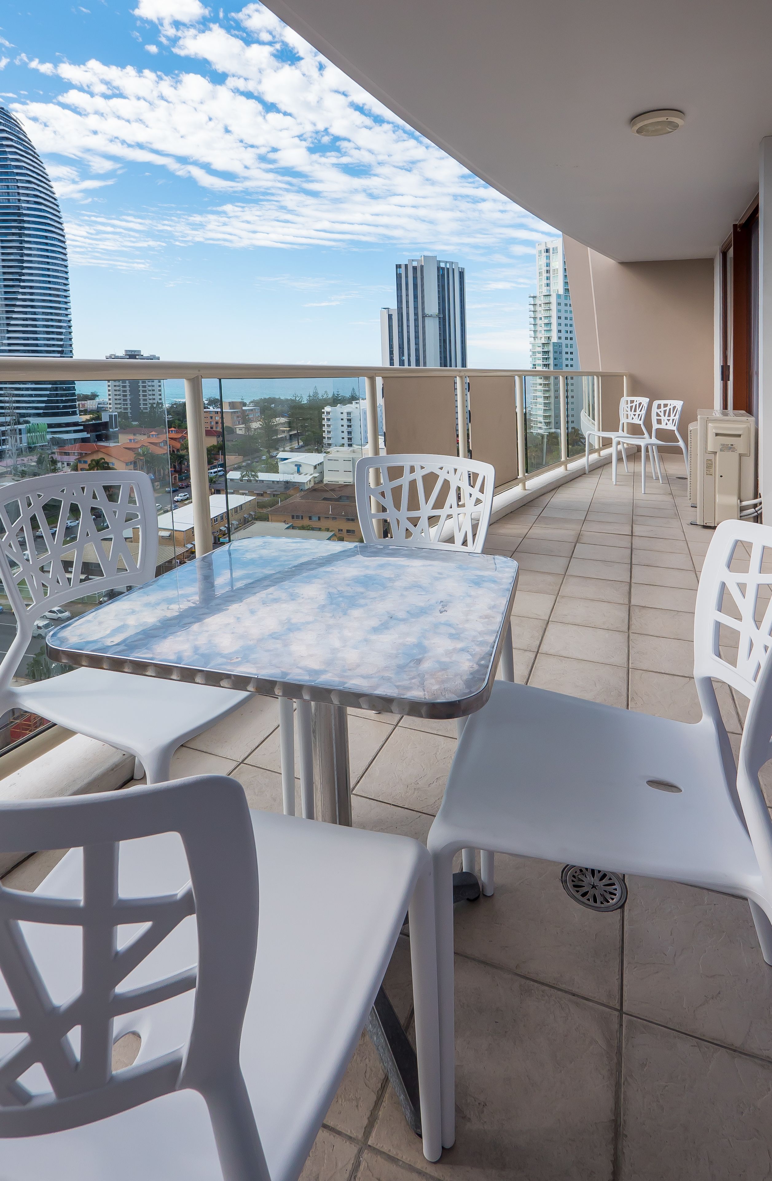 A balcony with white plastic chairs and a table, overlooking a cityscape with tall buildings.
