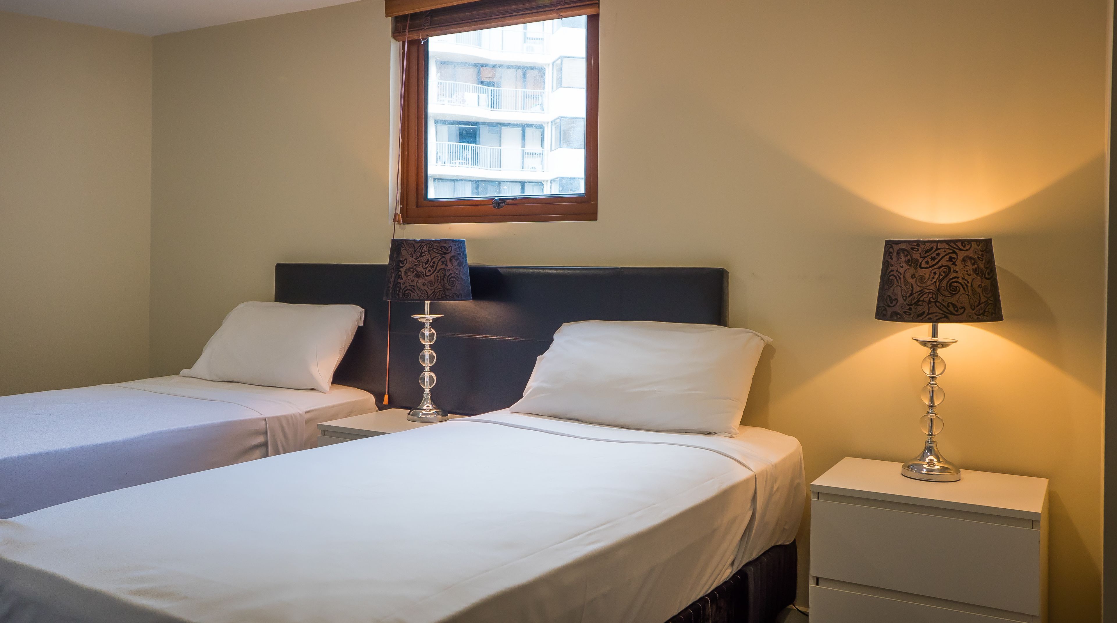 A hotel room with two single beds, a window with a view of buildings, and two bedside lamps.