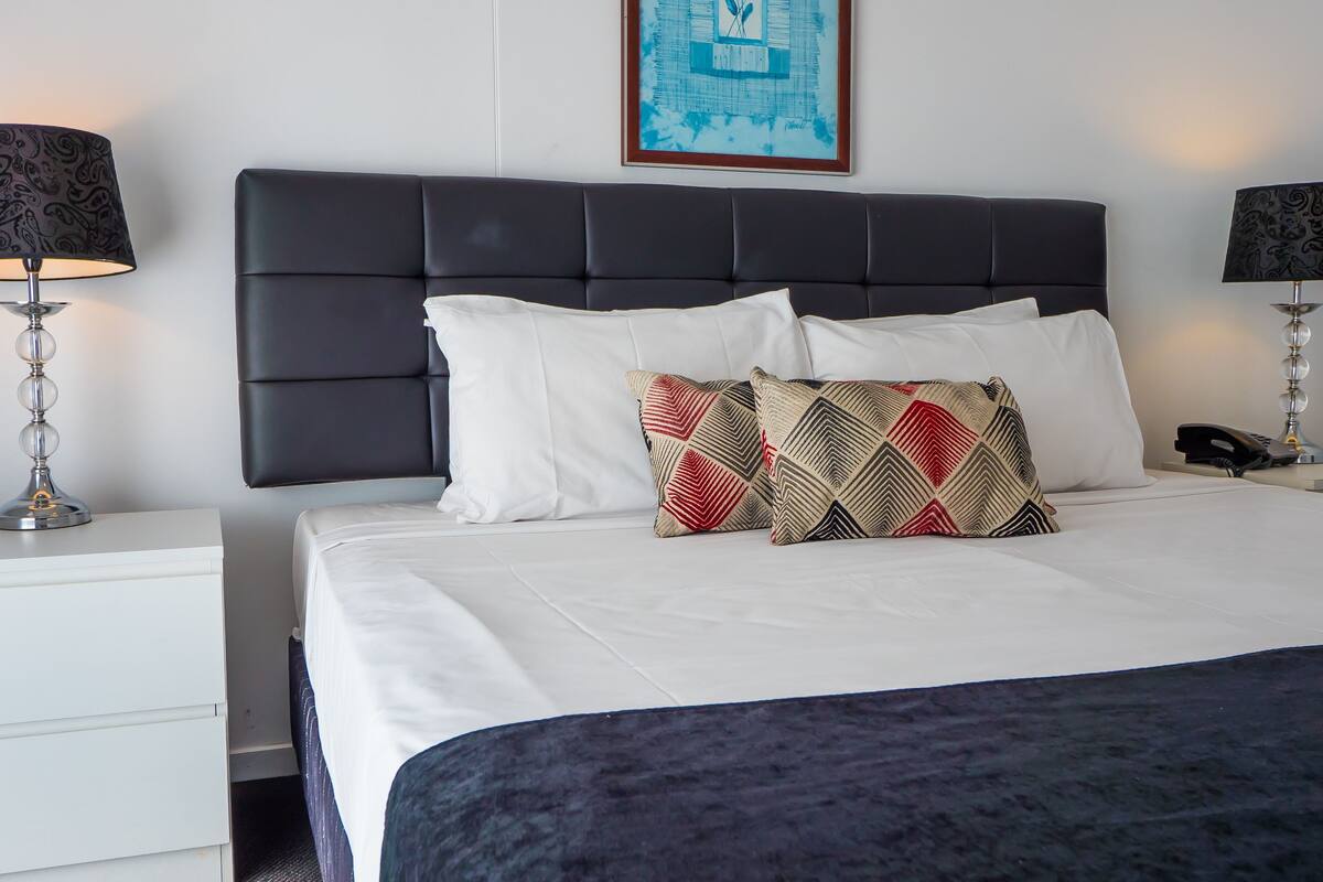 Een net opgemaakt bed met witte lakens, een donker hoofdbord en decoratieve kussens. Er staan twee nachtkastjes met lampen en er hangt een lijst met kunst aan de muur.
