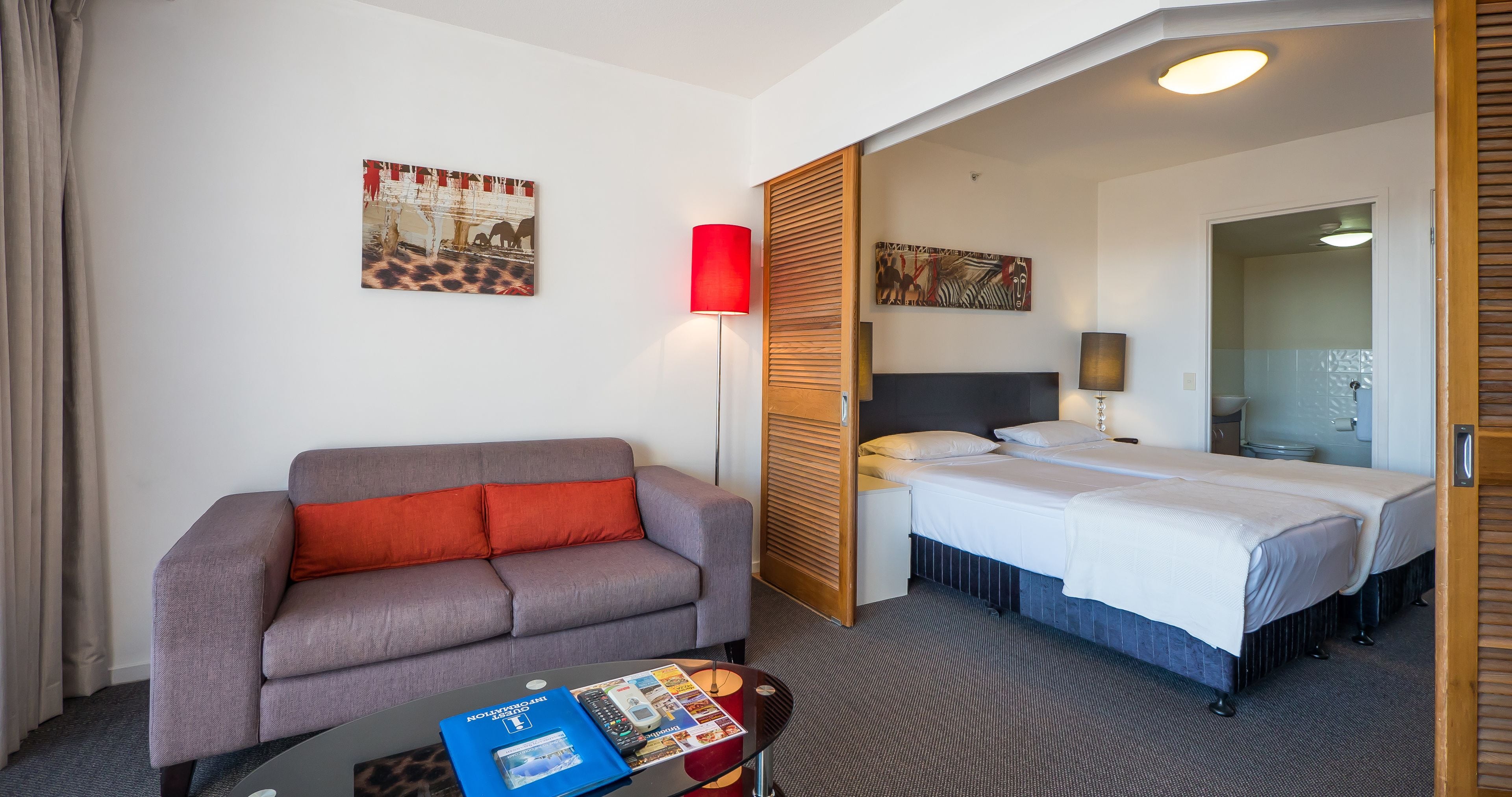 A hotel room with a grey sofa, a bed with white bedding, a red lamp, and a small table with a magazine and remote control.