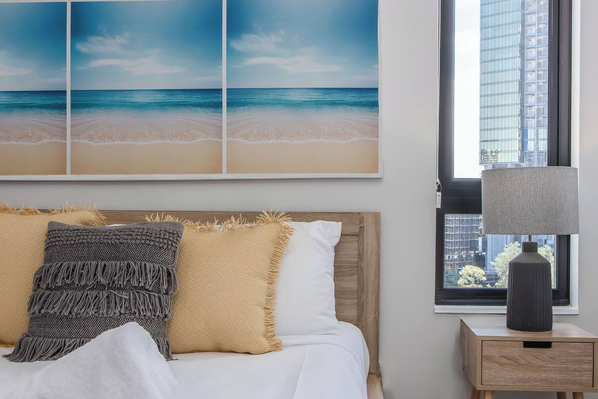 Une chambre à coucher avec un lit, des oreillers, une table de chevet, une lampe et une grande fenêtre donnant sur un immeuble.