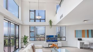 Ein geräumiges Wohnzimmer mit einem weißen Sofa, einem Glastisch und Blick auf eine Stadtlandschaft durch große Fenster.