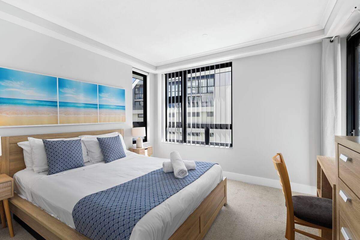 Une chambre d’hôtel moderne avec un grand lit, une table de chevet en bois, un bureau avec des tiroirs et une fenêtre équipée de stores.