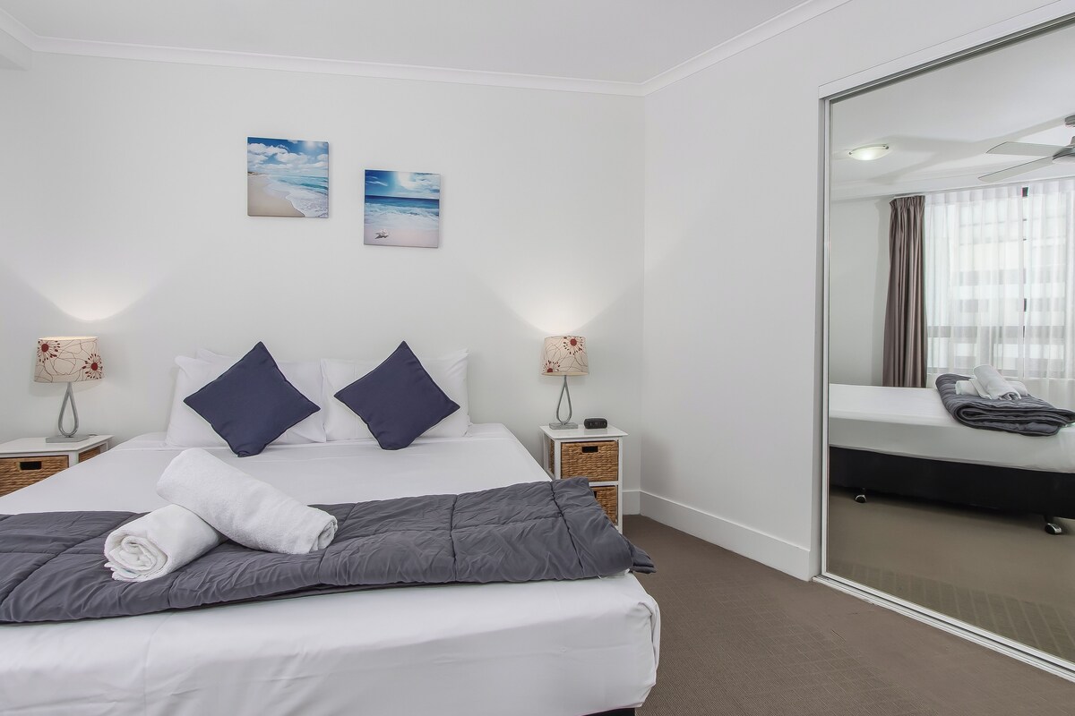 Habitación de hotel con una cama bien hecha, dos lámparas fijadas a la pared y un espejo grande que refleja un baño.
