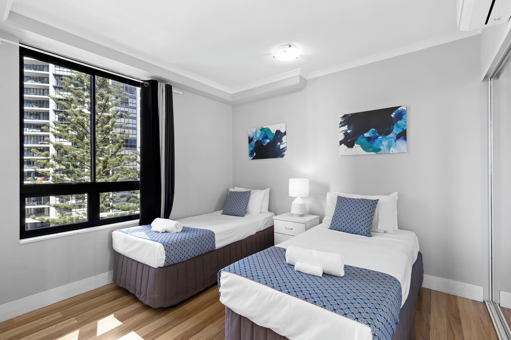 A hotel room with two beds, a window with curtains, a nightstand, and artwork on the wall.