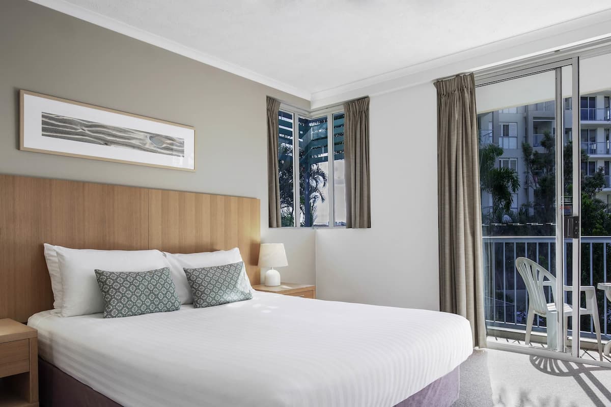 A hotel room with a large bed, a nightstand, a bedside table, a lamp, a chair, and a balcony with a view of buildings and trees.