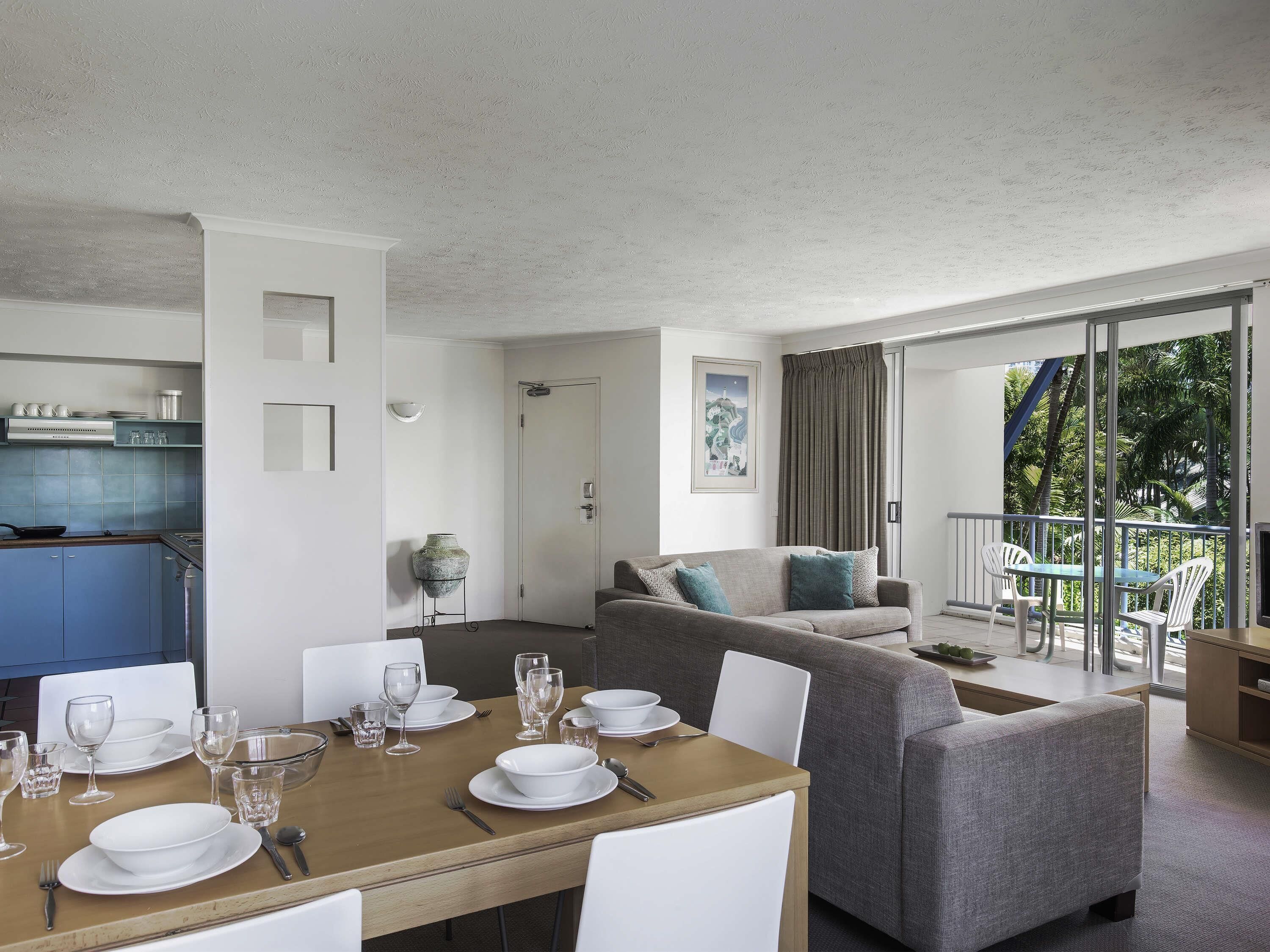 A modern living room with a dining area, a kitchen, and a balcony with a view of greenery.
